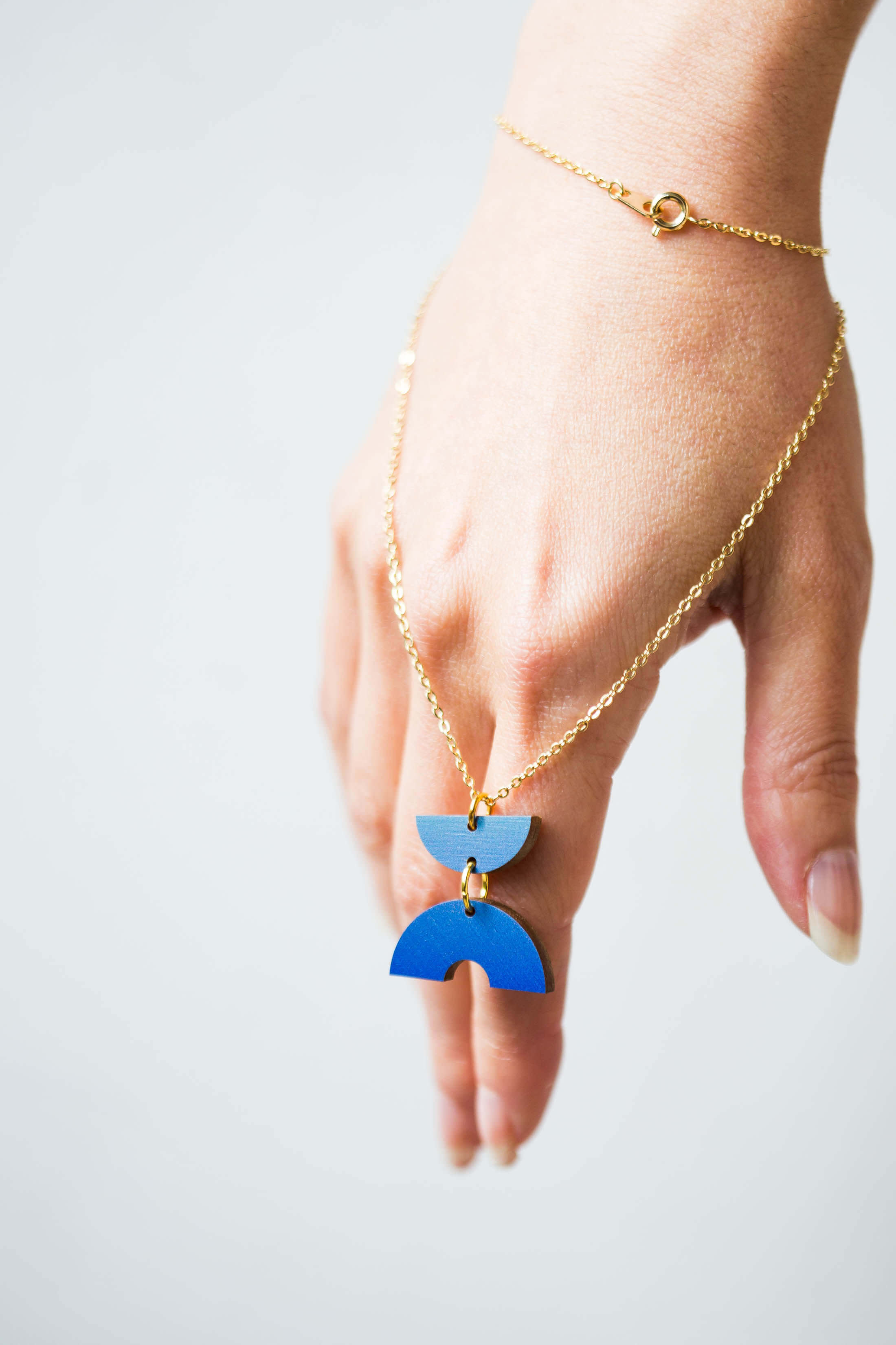Gold chain necklace with a blue, geometric, wooden pendant featuring two stacked semicircle shapes.