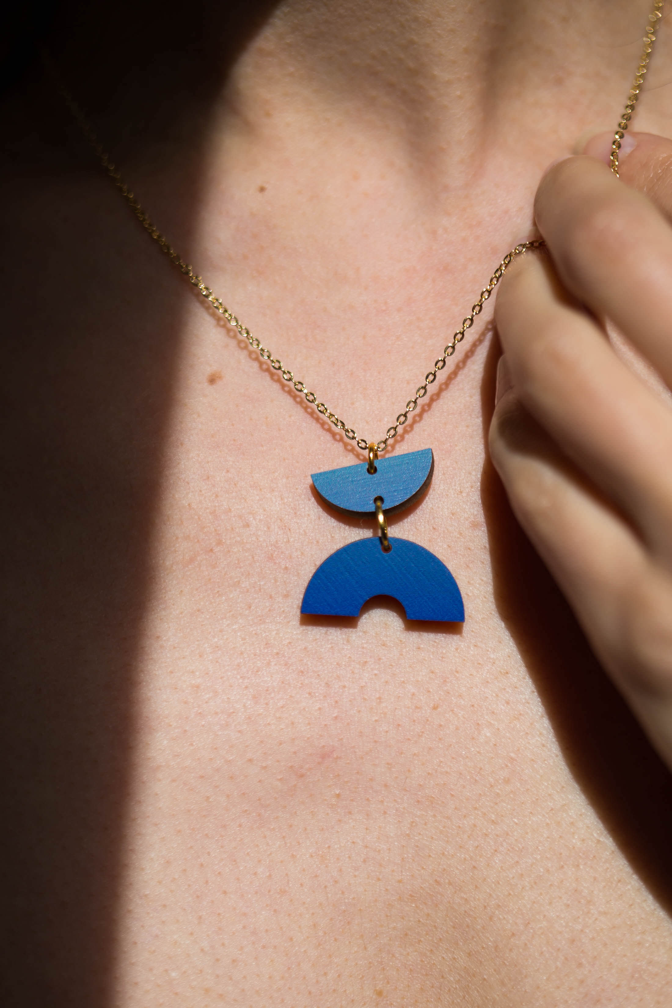Gold chain necklace with a blue, geometric wooden pendant featuring two stacked semicircle shapes.