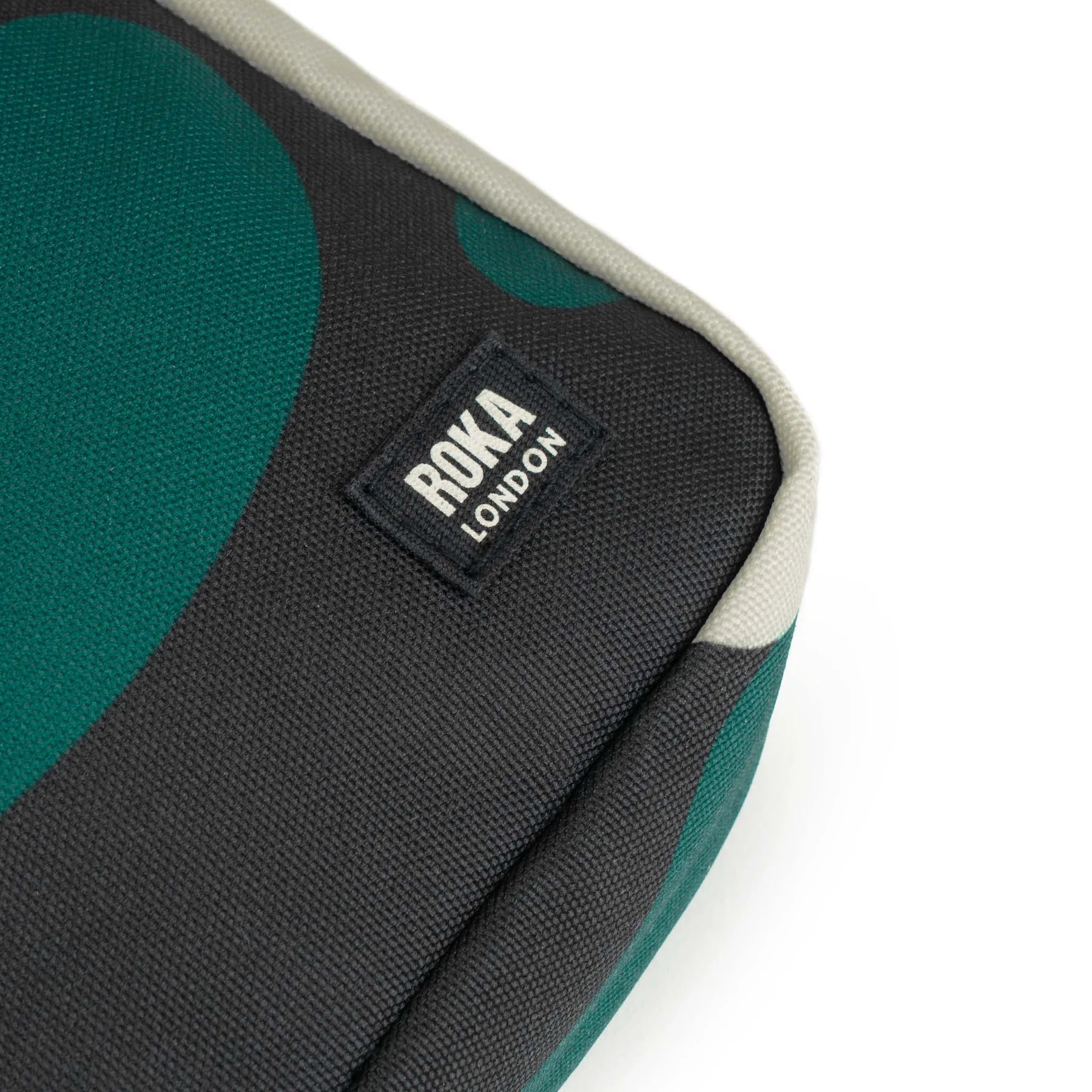 Close-up of a dark gray canvas bag with teal abstract shapes and a "ROKA LONDON" label. The bag has a light beige trim.