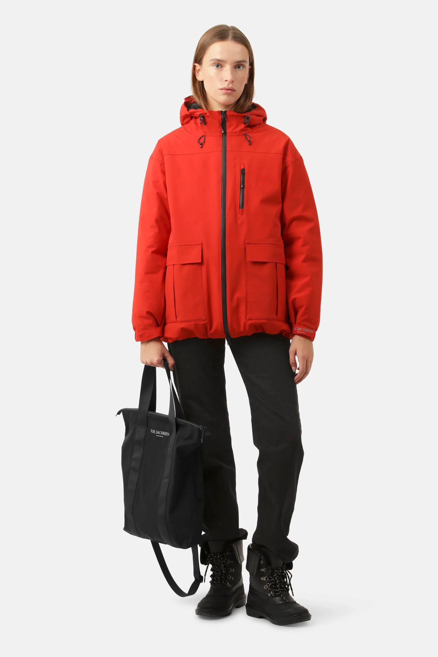 Woman in red hooded jacket, black pants, and boots, carrying a black Ilse Jacobsen tote bag.