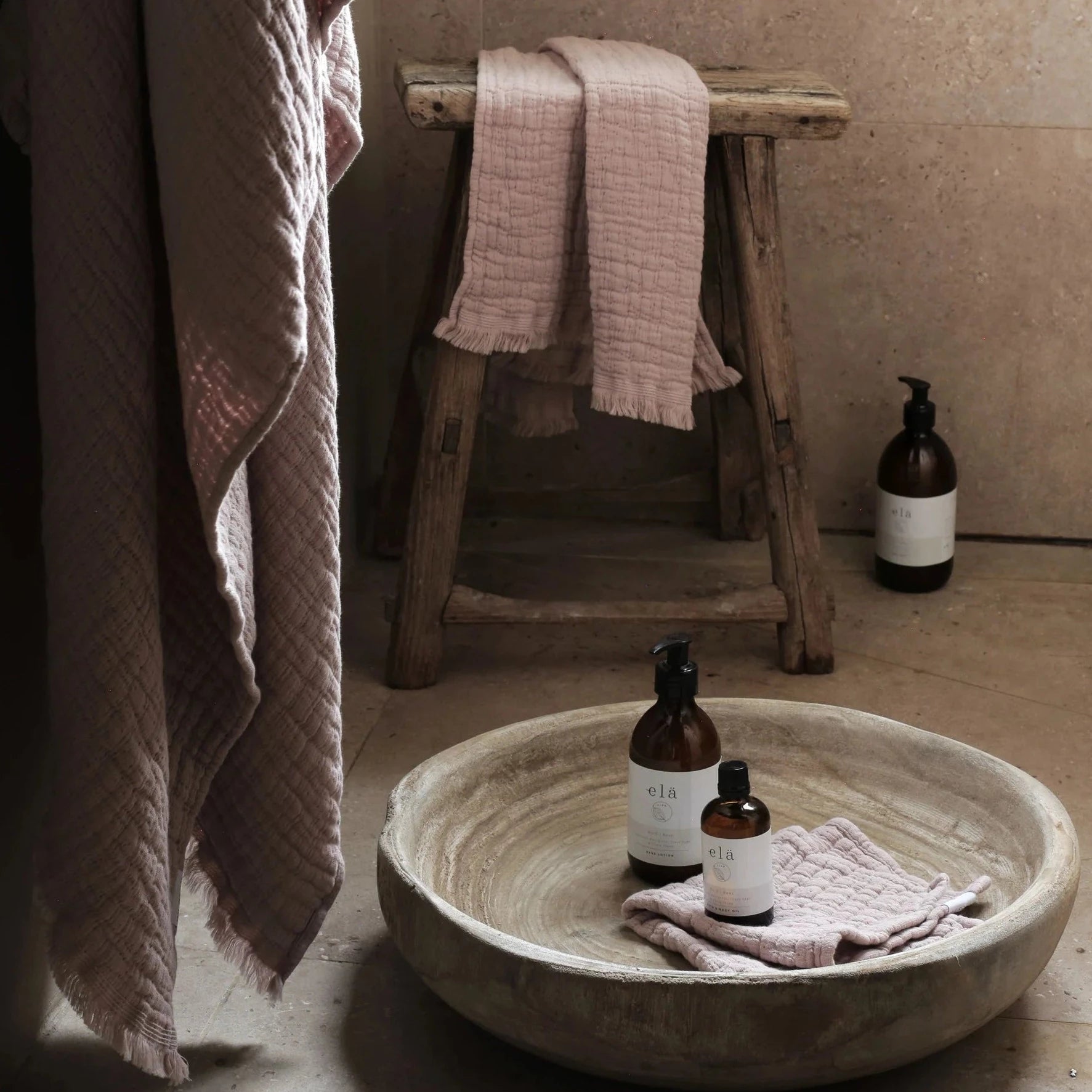 Large, rustic wooden bowl containing two amber bottles of elá body care products and a pale pink textured washcloth.