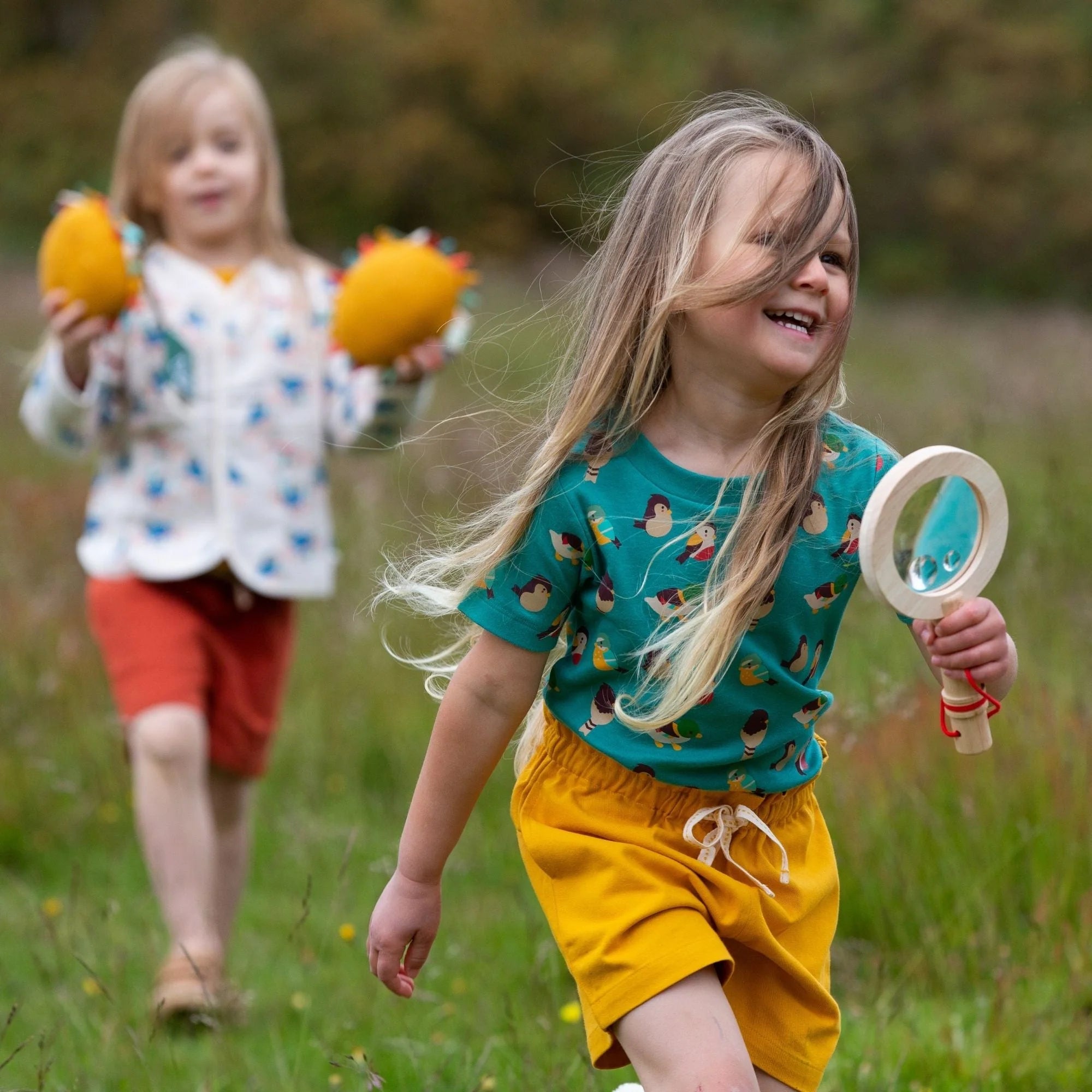 A girl holds a wooden magnifying glass; she wears a teal bird-print shirt and mustard yellow shorts.