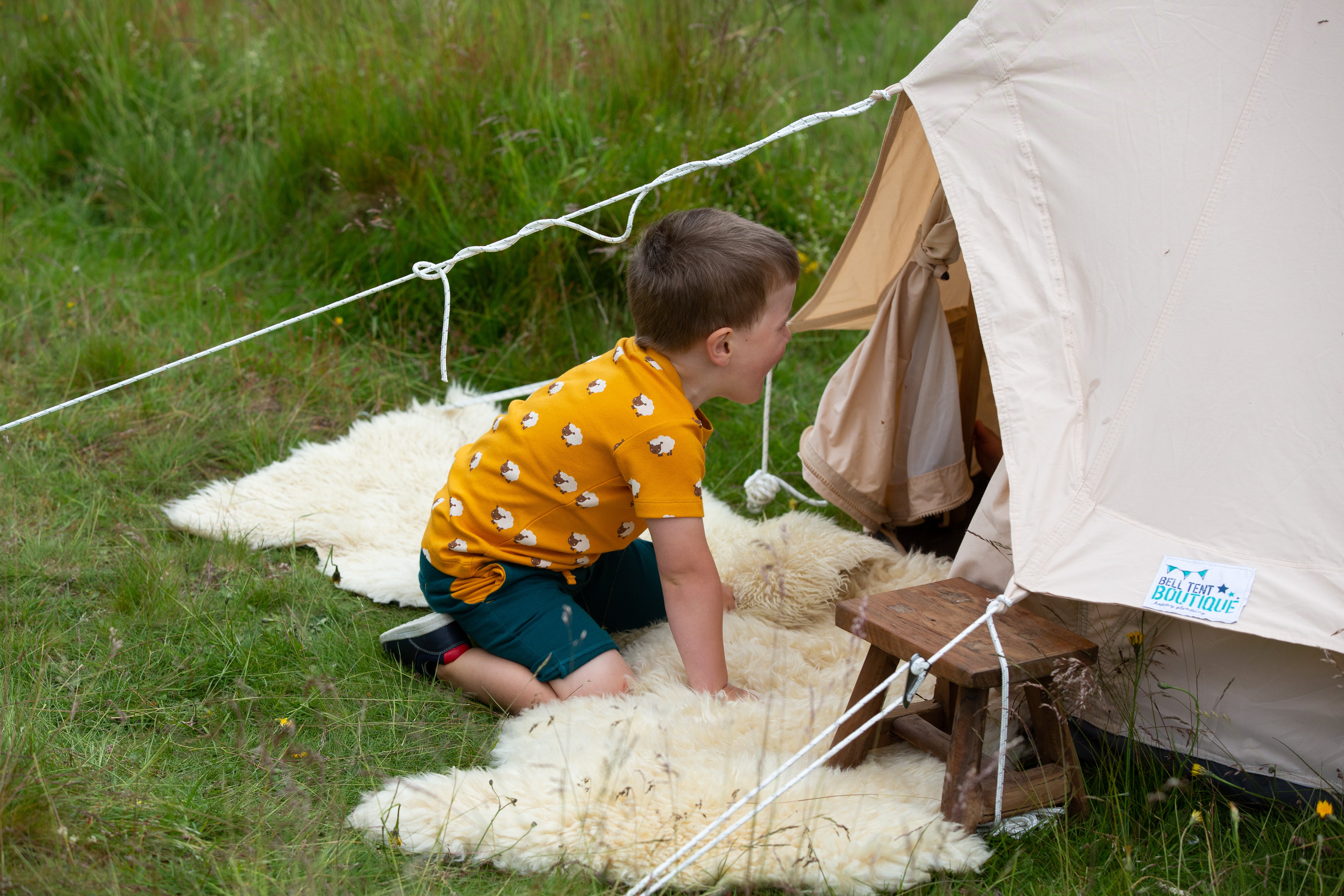 Cream-colored bell tent with a small wooden stool and child in front.  The tent has a logo that reads "Bell Tent Boutique".