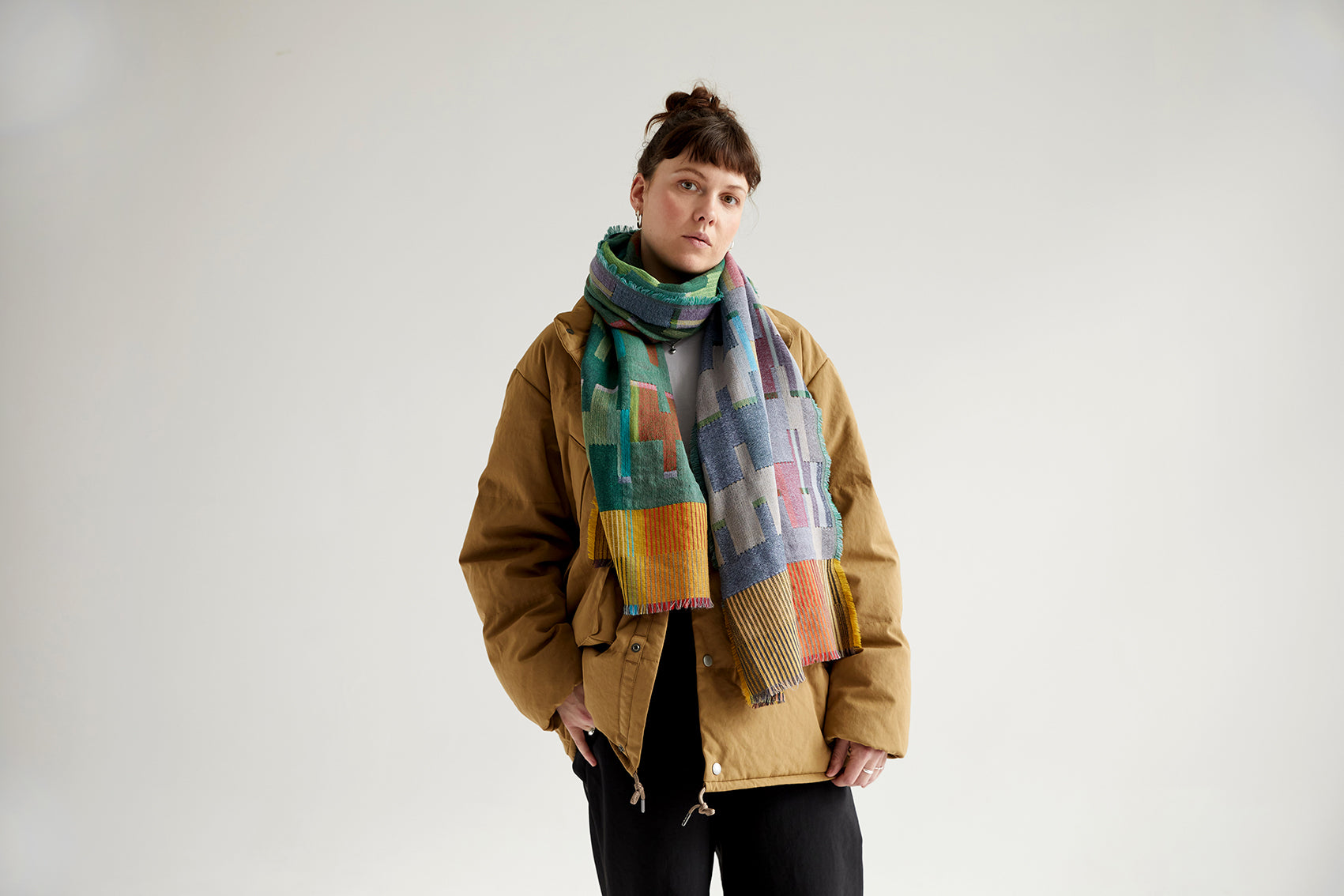A multicolored geometric patterned scarf in teal, orange, and gray, draped around a person wearing a tan jacket.