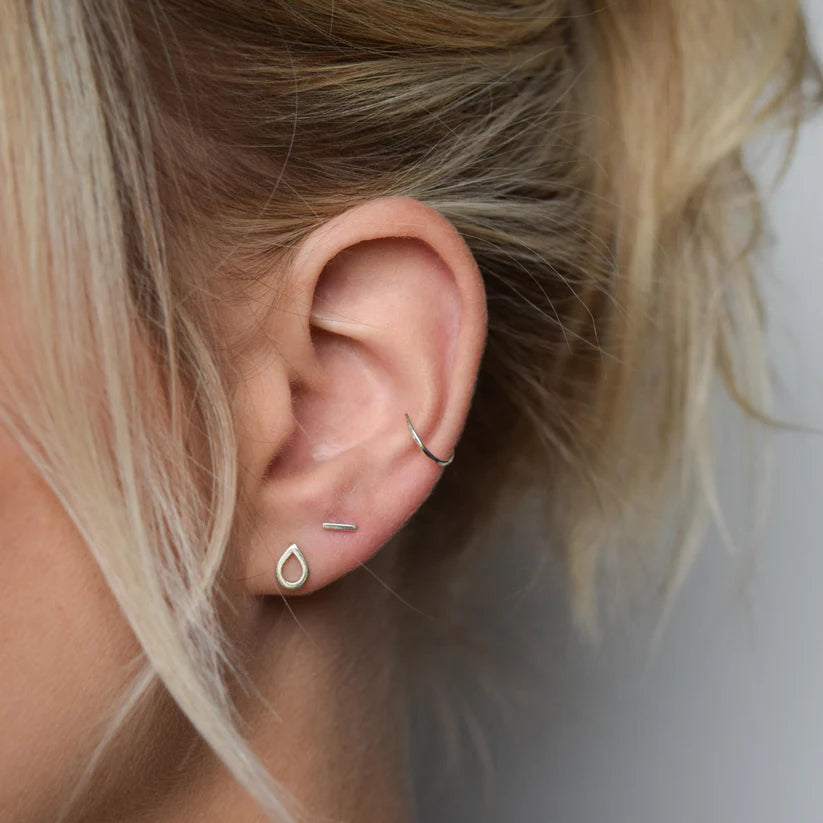 Close-up of an ear with three silver earrings: a teardrop stud, a small bar stud, and a small hoop in the upper cartilage.