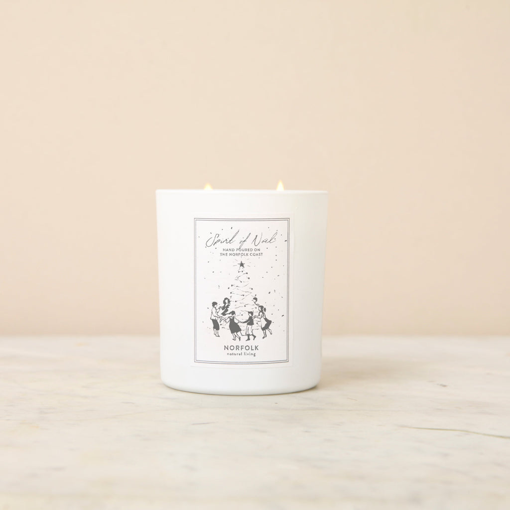 a double wicked, white jar candle, set on a white marble surface against a peach background, featuring a black and white label stating 'Norfolk Natural Living - Spirit of Noel' and depicting children, holding hands, dancing around a decorated Christmas tree