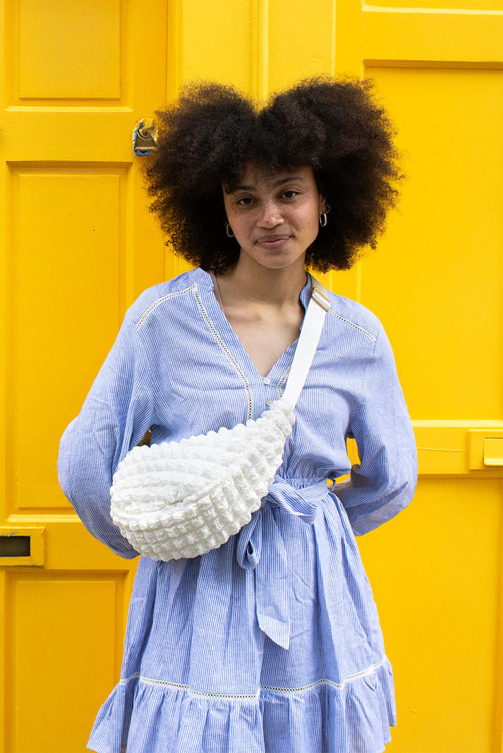 White textured crescent-shaped fanny pack worn by a person in a blue and white striped dress.