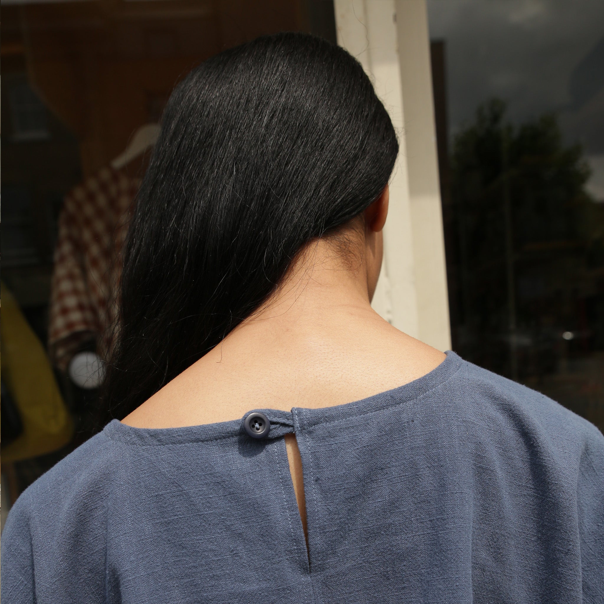 Rear view of a slate blue linen top with a keyhole back and single button closure.  Long black hair is visible.