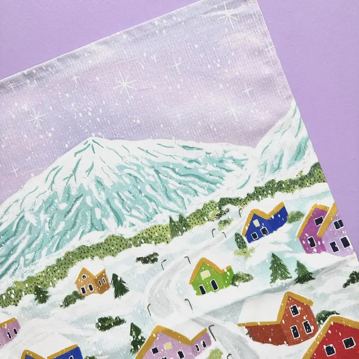 Snowy winter village scene tea towel; colorful houses nestled in a snow-covered landscape beneath a large, snow-capped