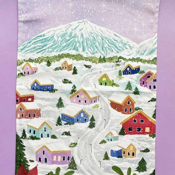 Winter village scene garden flag; colorful houses covered in snow, winding road, and snow-capped mountain in the background.