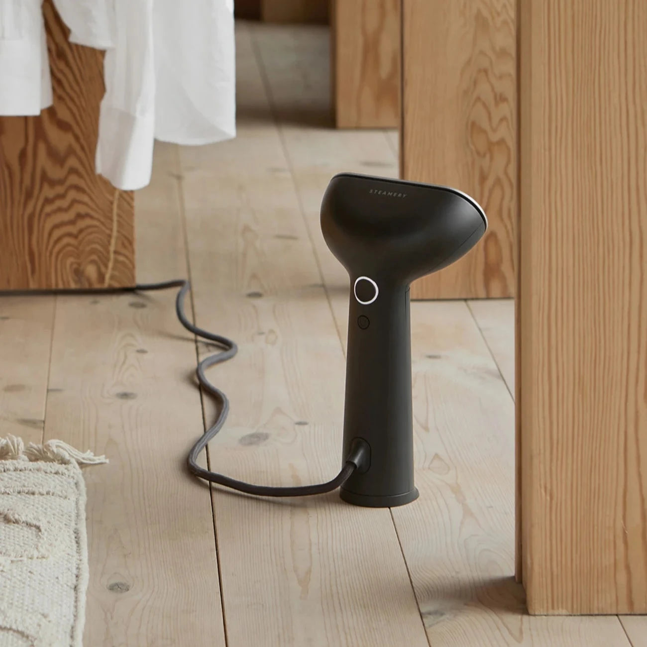 Black Streamery handheld garment steamer on light wood floor.  Cord plugged in.