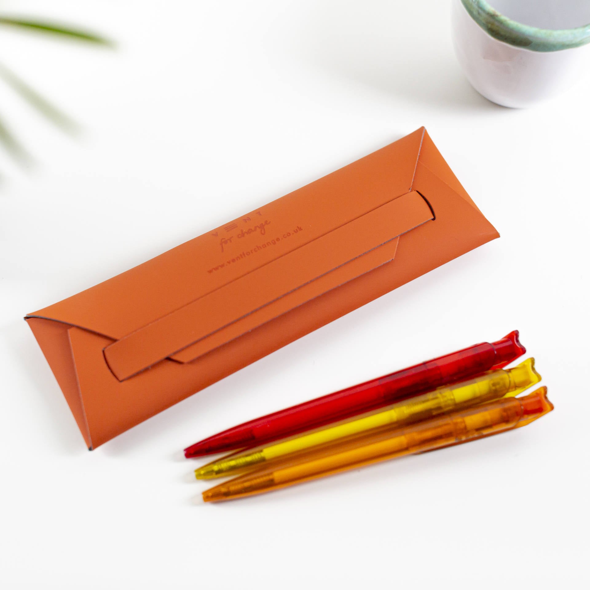 Burnt orange leather pen case with subtle branding, accompanied by three translucent pens in red, yellow, and orange.