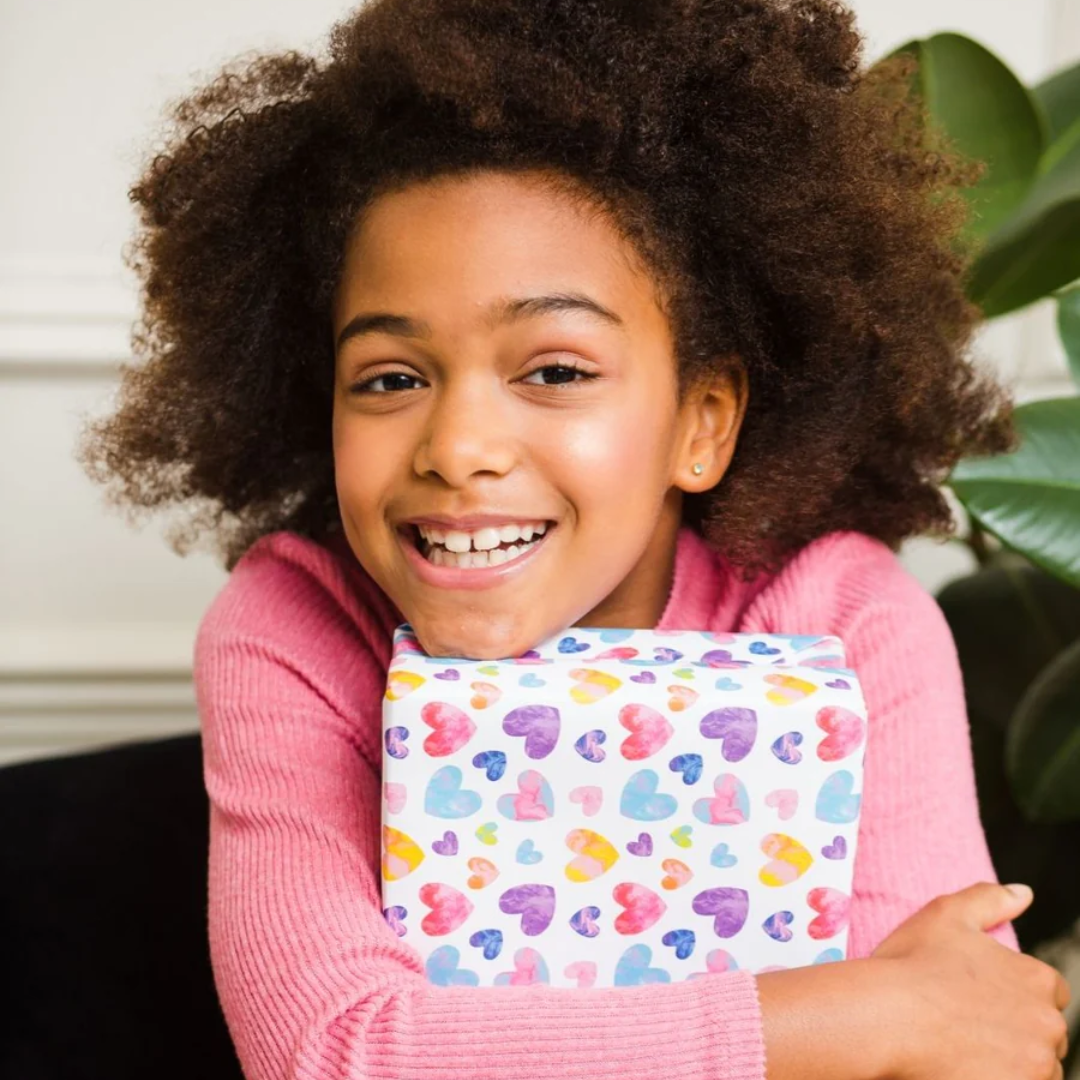 Smiling girl hugging a gift wrapped in colorful heart-patterned paper.