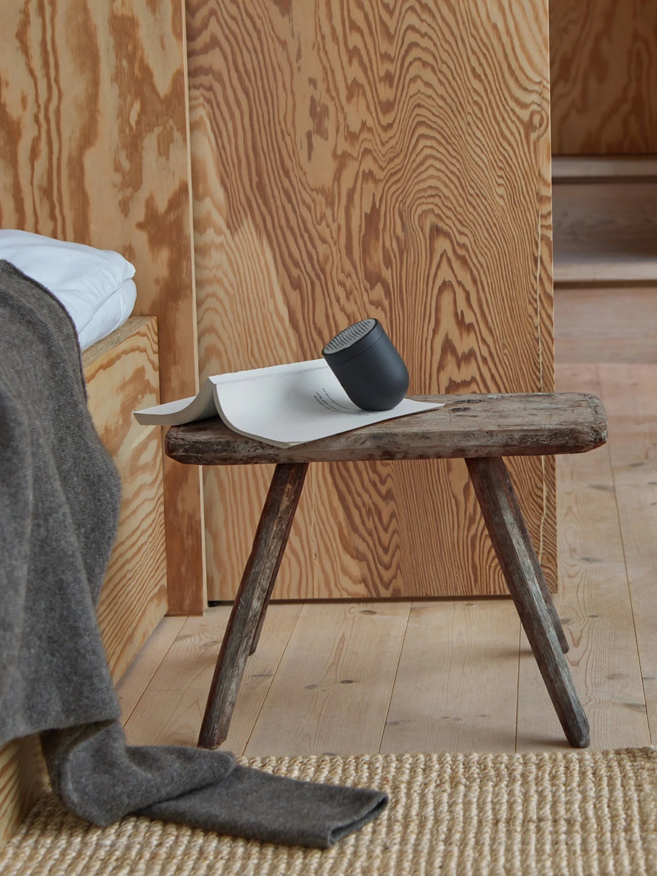 Dark gray cylindrical speaker resting on a small wooden stool, next to a folded piece of paper.
