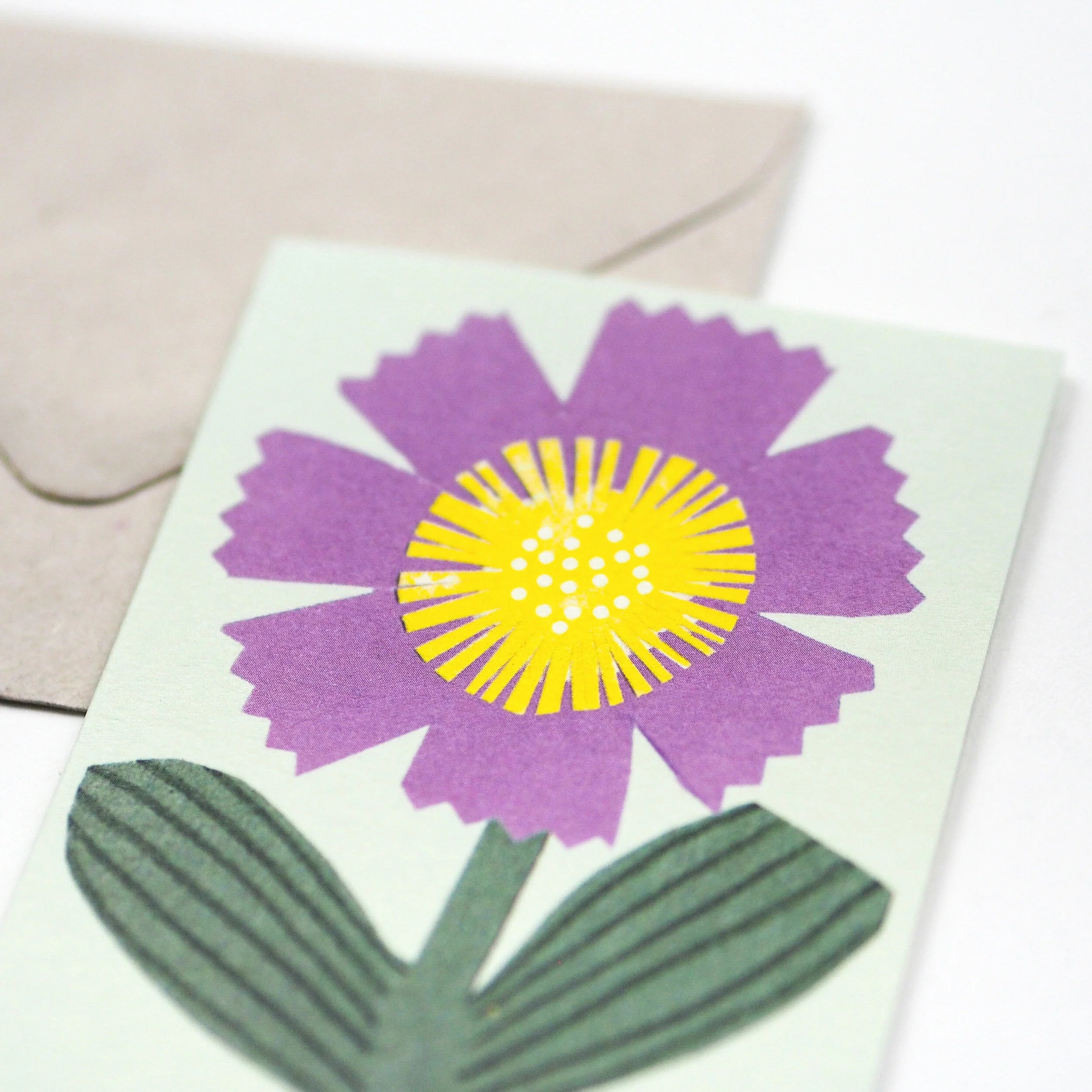 Close-up of a greeting card featuring a stylized purple flower with a yellow center and green leaves on a light green