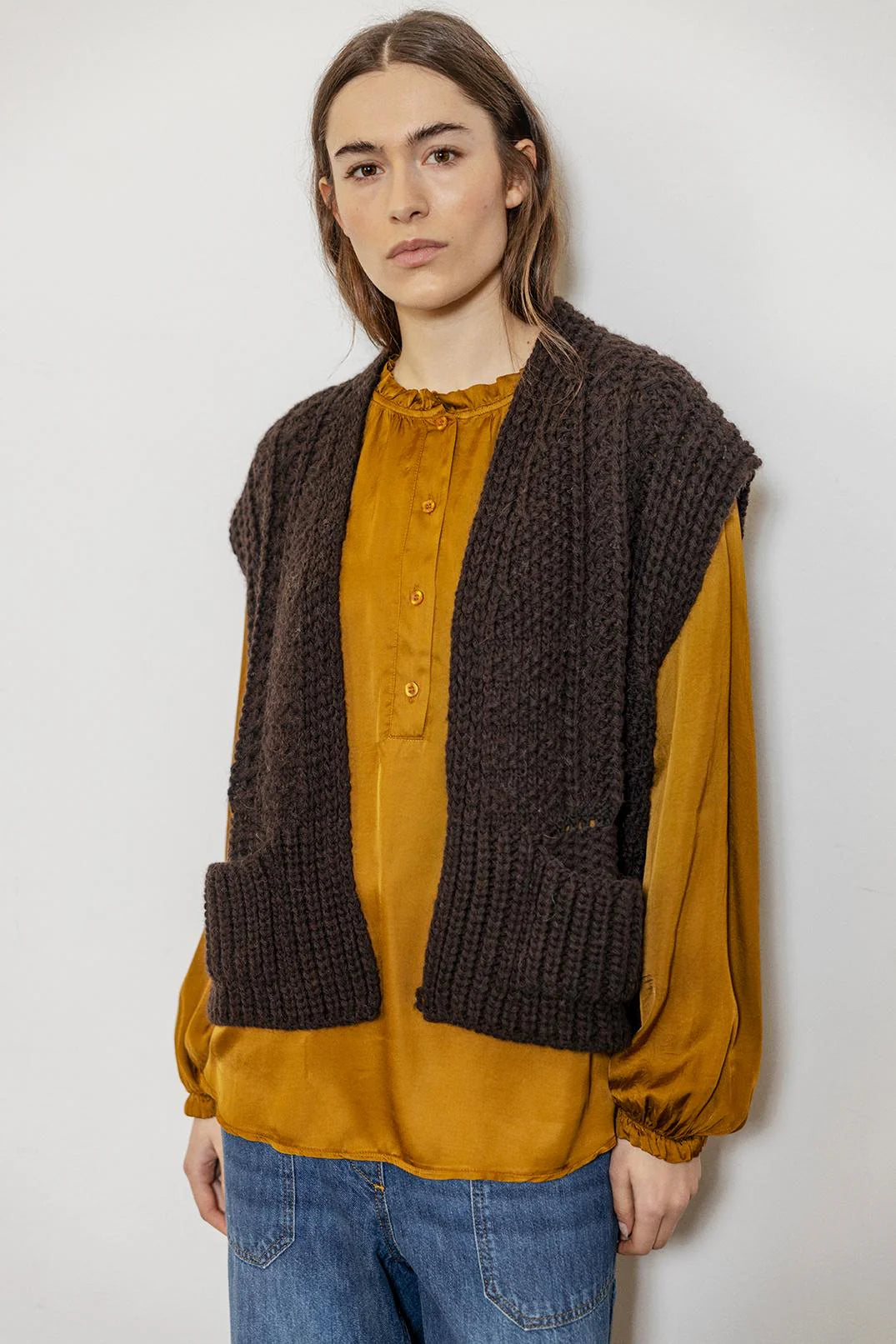 Person wearing a brown knitted vest over a mustard yellow shirt with blue jeans against a white background