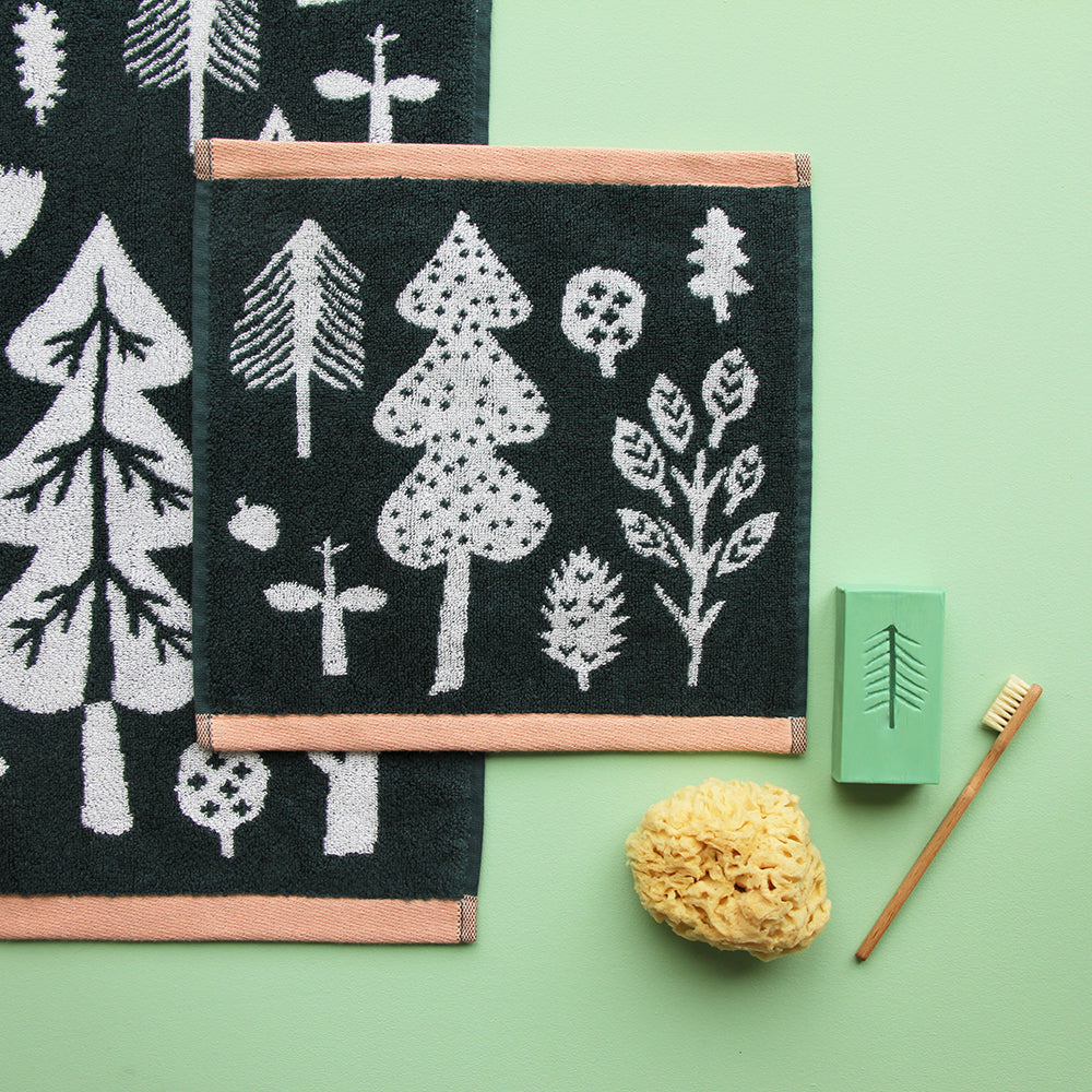 Two dark green towels with white tree and leaf designs, trimmed in peach, by Donna Wilson. Accompanied by a sponge and brush.