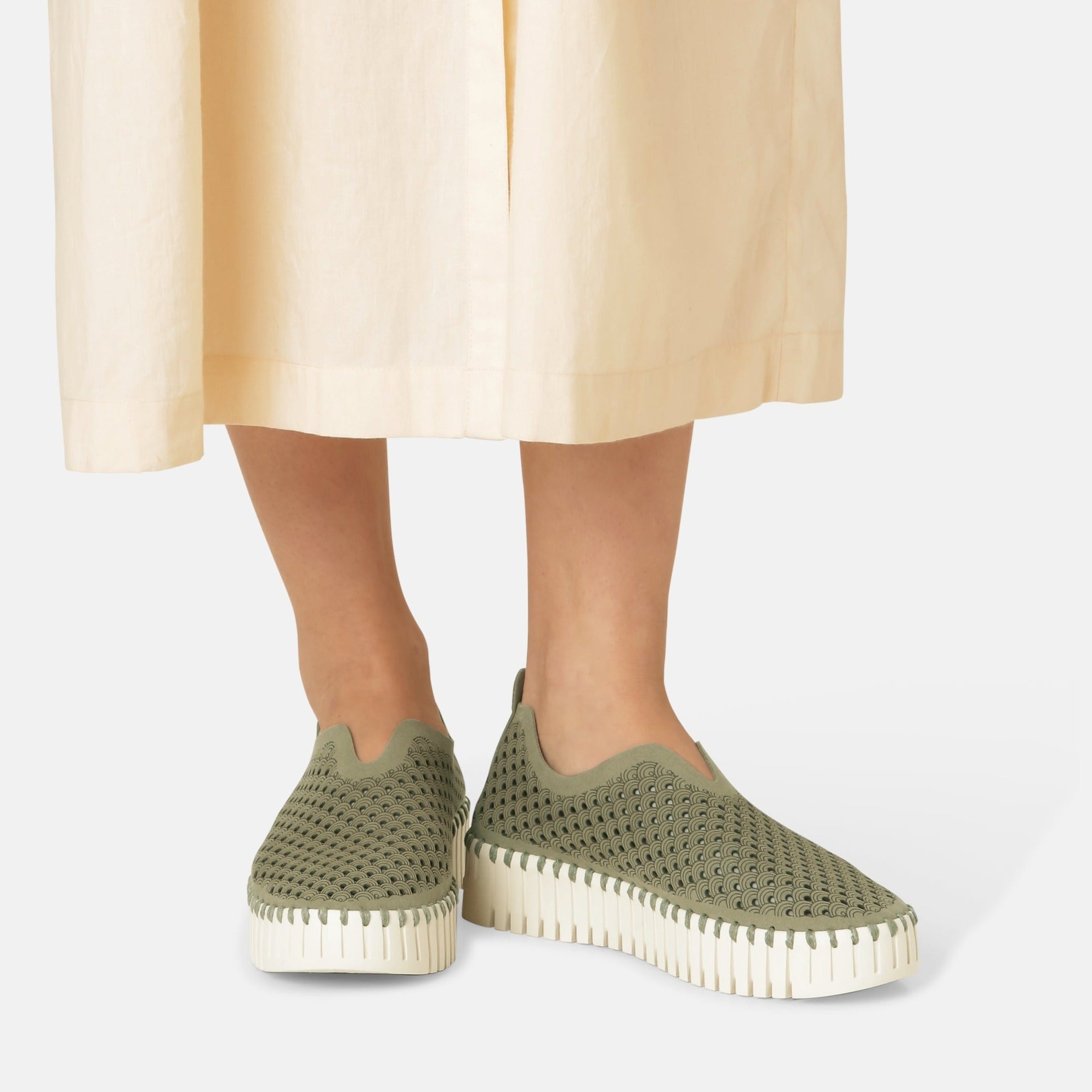 Olive green perforated slip-on shoes with a cream platform sole, worn by a model in a beige skirt.