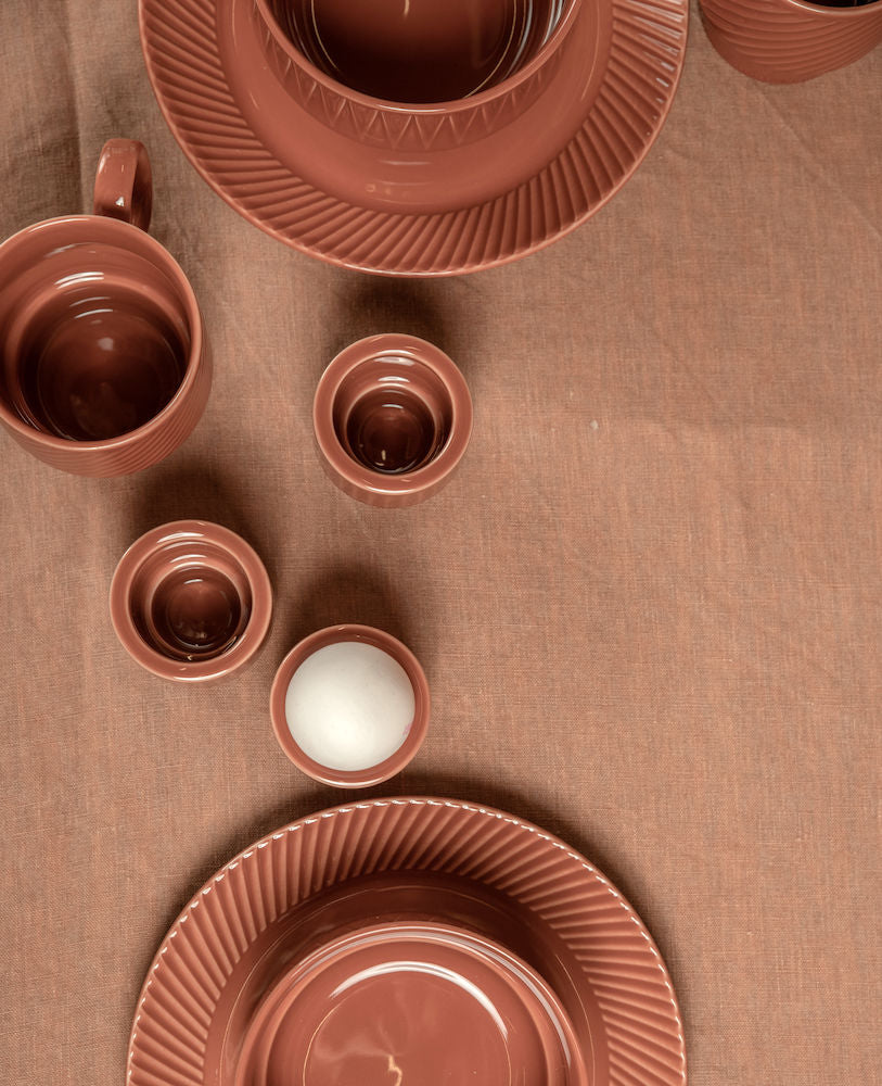 Set of terracotta-colored ceramic tableware: plates, bowls, mugs, and egg cups.  A single egg rests in one cup.
