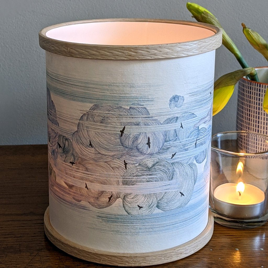Cylindrical candle lantern with a light wood base and top, featuring a blue and white illustration of clouds and birds.
