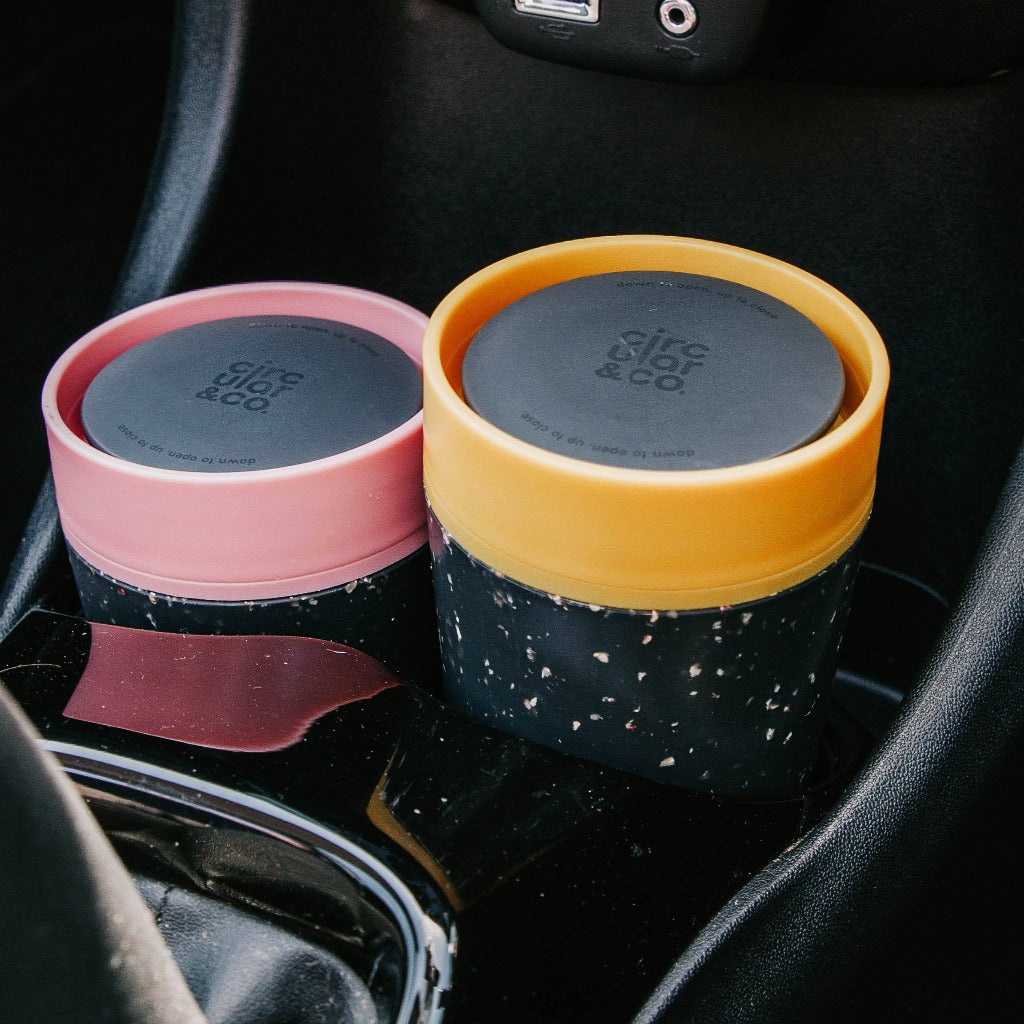Two Circulor & Co. reusable coffee cups; one pink, one mustard yellow, both with dark speckled bodies and lids.