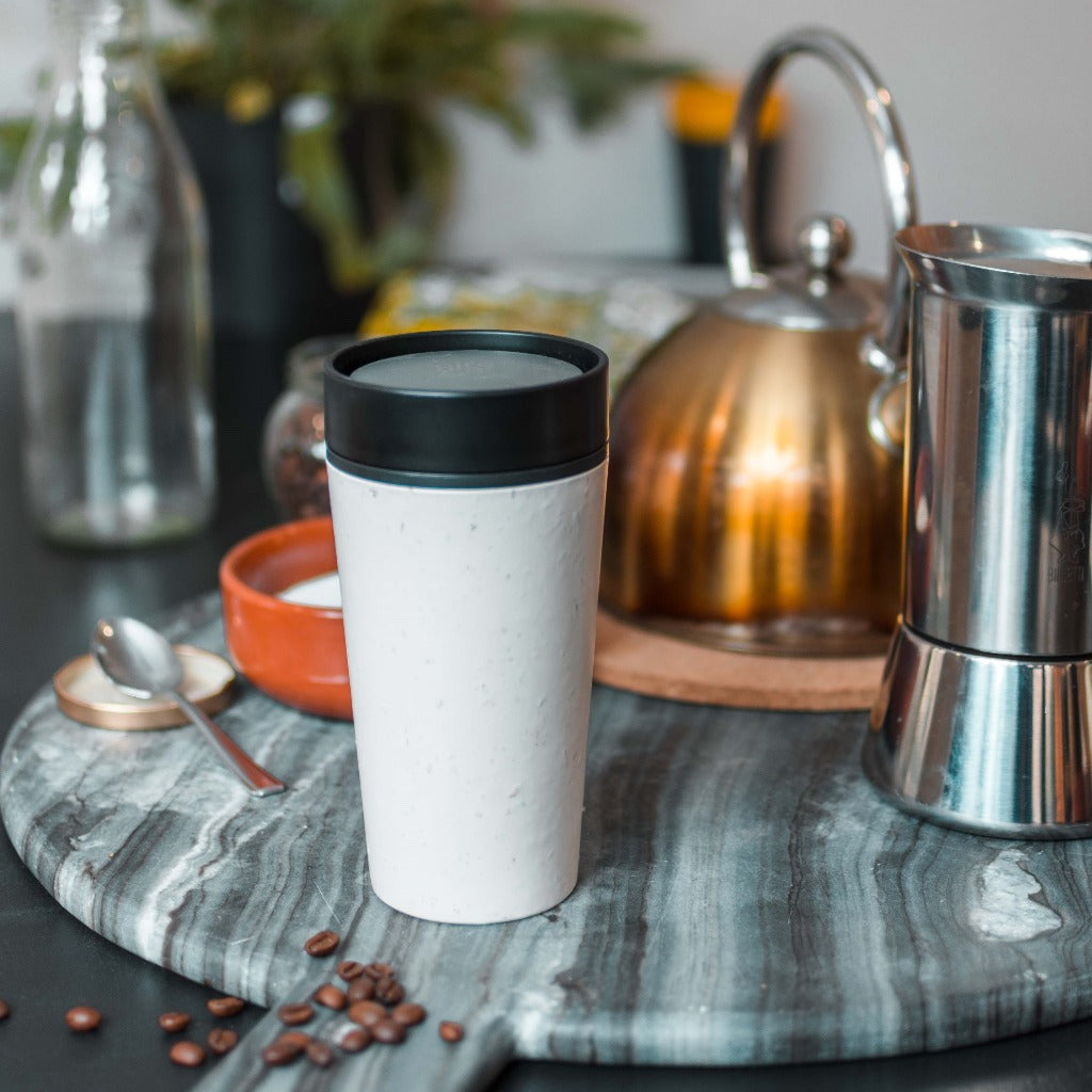 White reusable coffee cup with black lid, sitting on a marble surface.