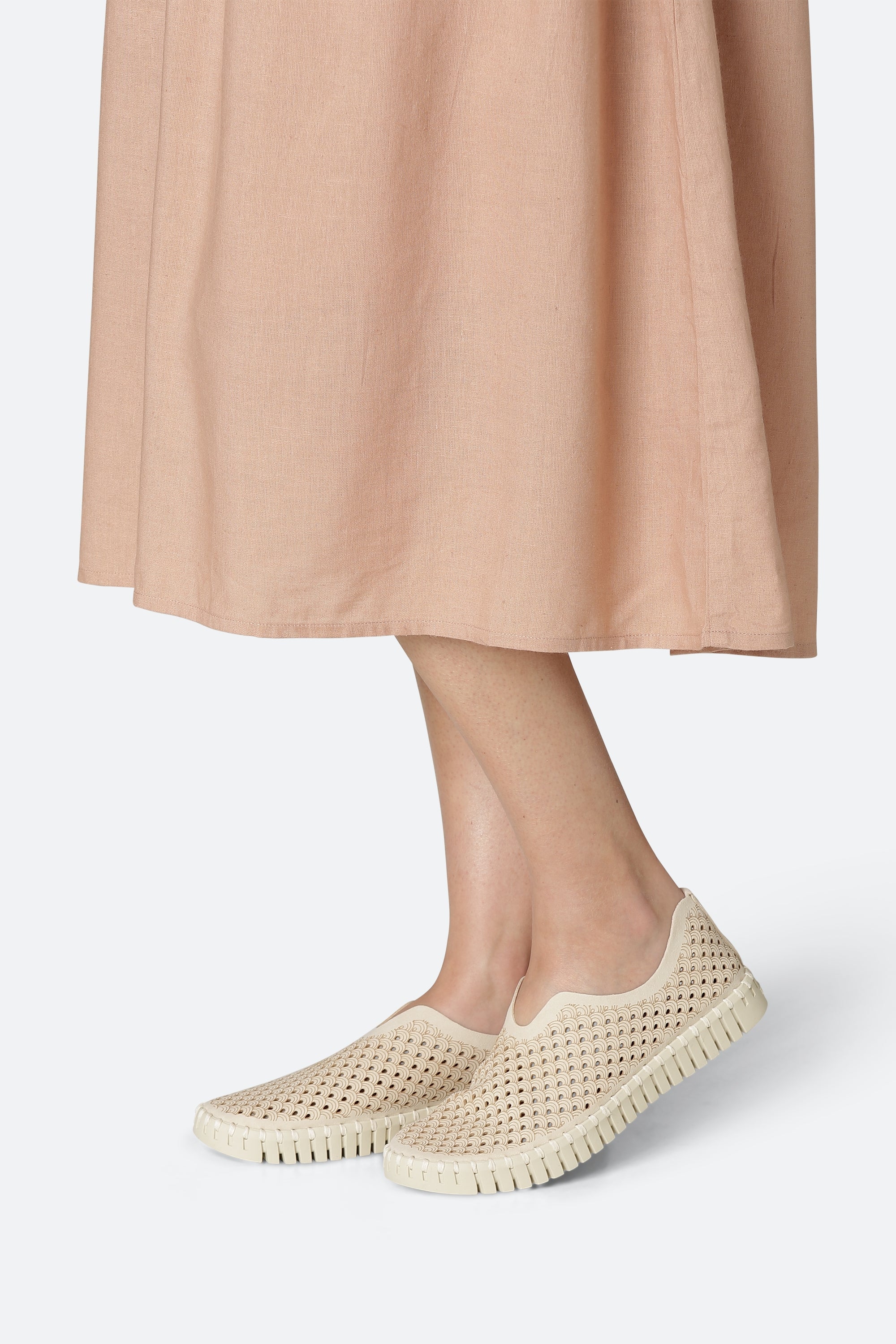 Beige slip-on shoes with a textured, perforated pattern on a cream-colored sole. The shoes are worn by a model whose legs and a