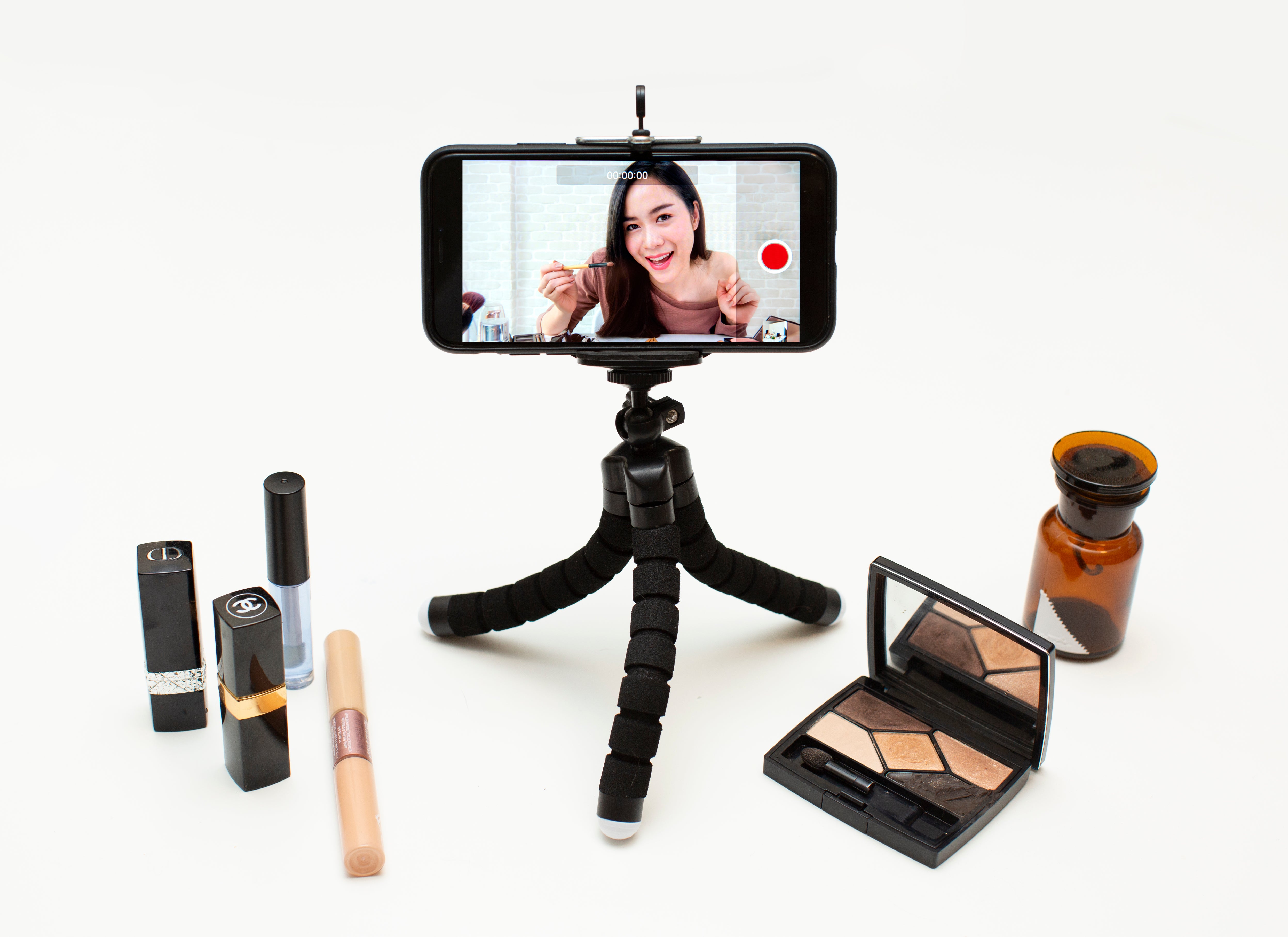 Smartphone on tripod displaying a beauty tutorial video;  surrounding it are lipsticks, eyeshadow palette, and concealer.