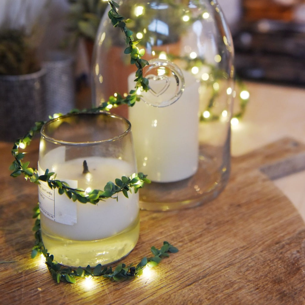 LED string lights with faux greenery wrapped around a candle and a glass cloche.