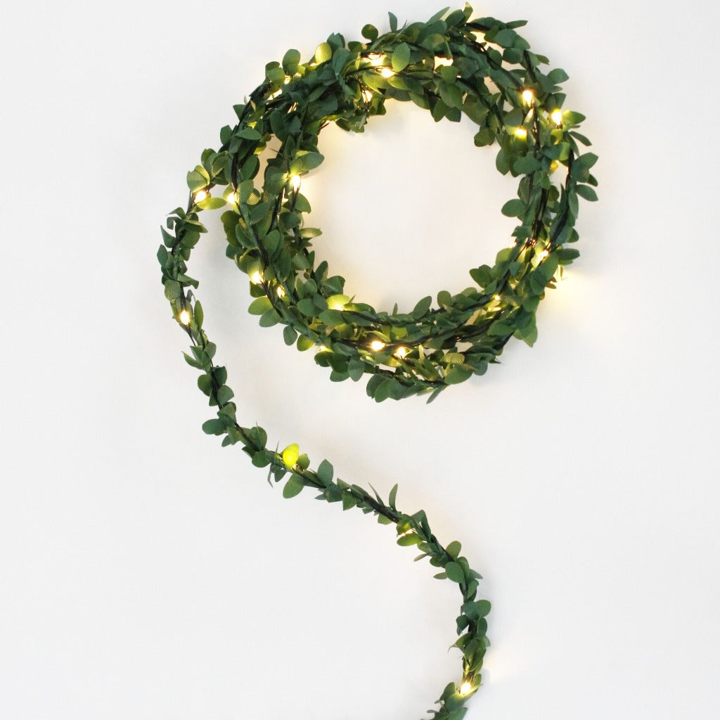 LED string lights woven into artificial greenery garland. Warm white lights illuminate the leaves.
