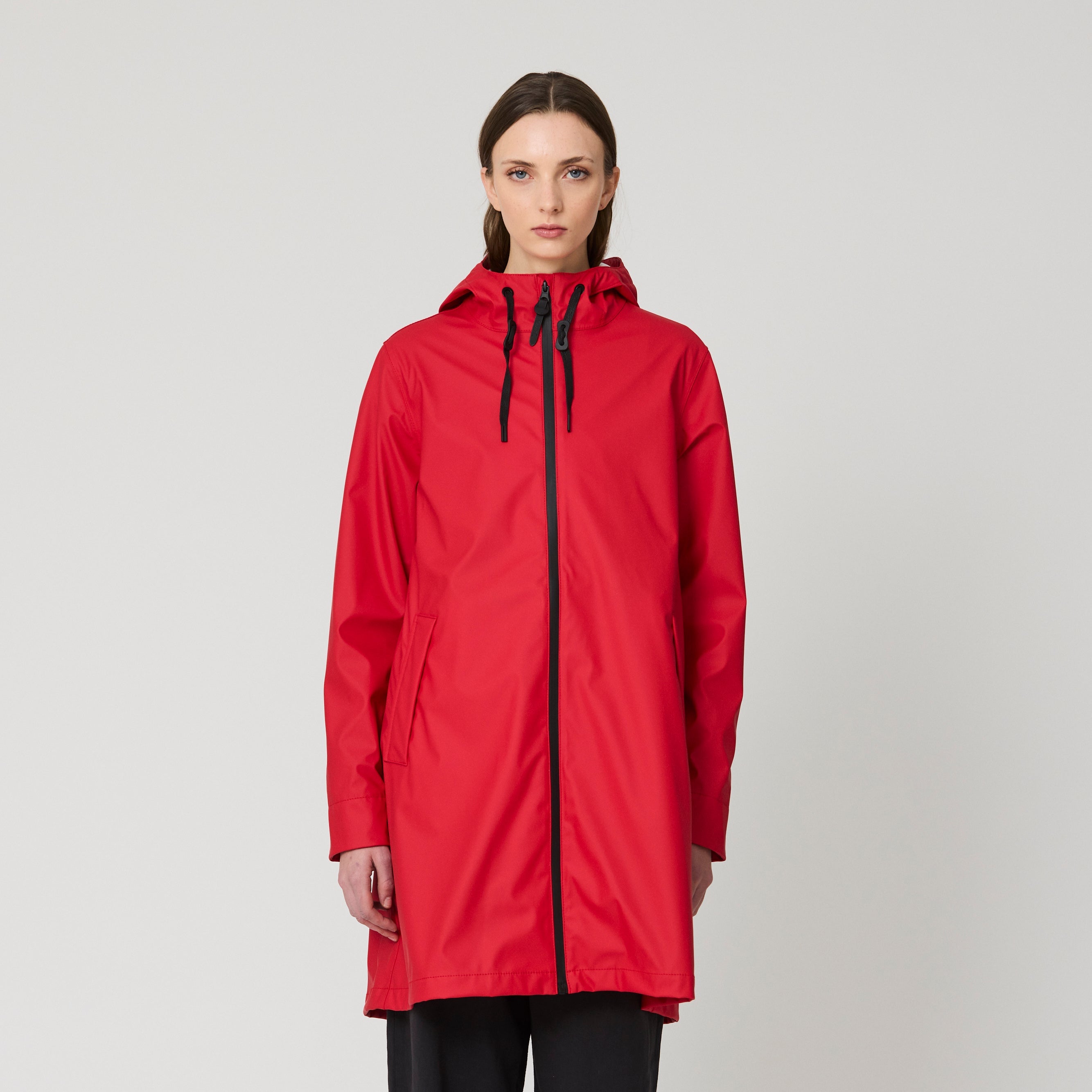 A model stands against a light gray studio background wearing a bright red hooded raincoat with a black front zipper and black drawstrings, paired with black pants and blue-and-black sneakers.