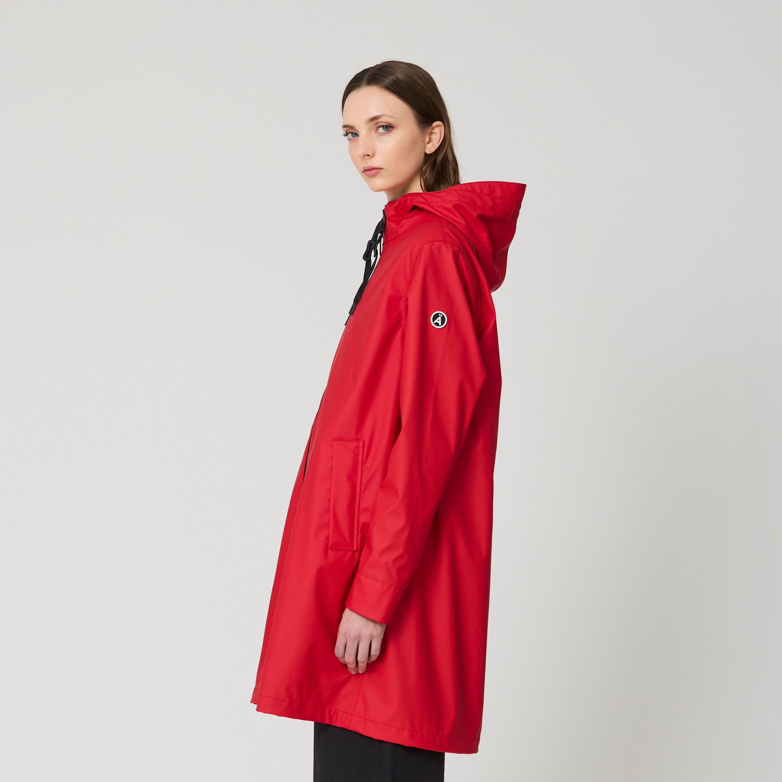 A woman modeling a bright red hooded raincoat, paired with black pants and blue sneakers, standing against a light gray studio background.