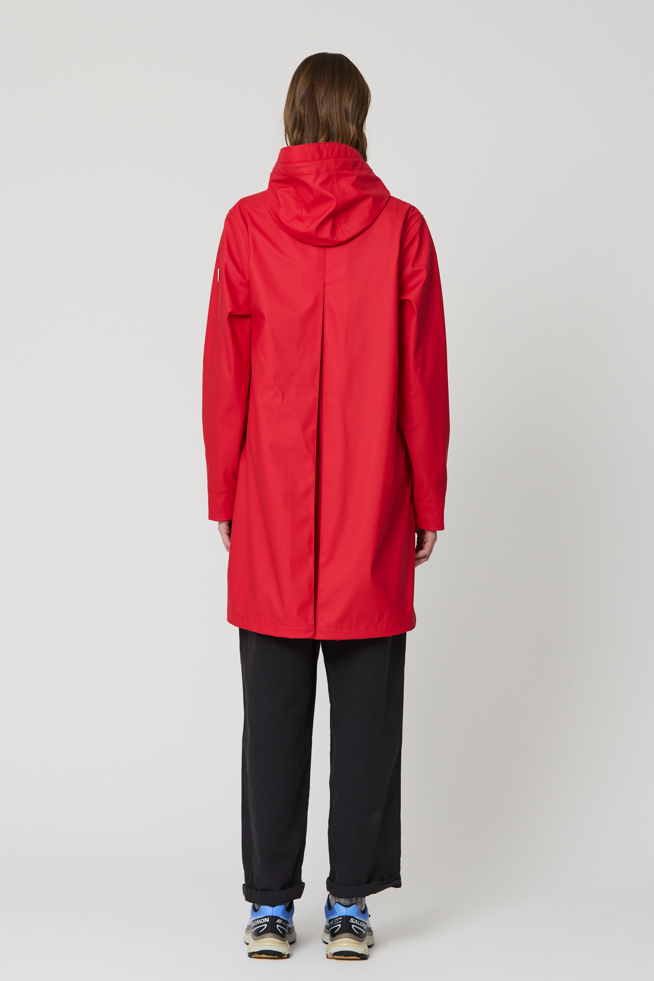 A model viewed from the back wearing a bright red hooded raincoat, black wide-leg trousers, and blue sneakers against a light gray studio background.