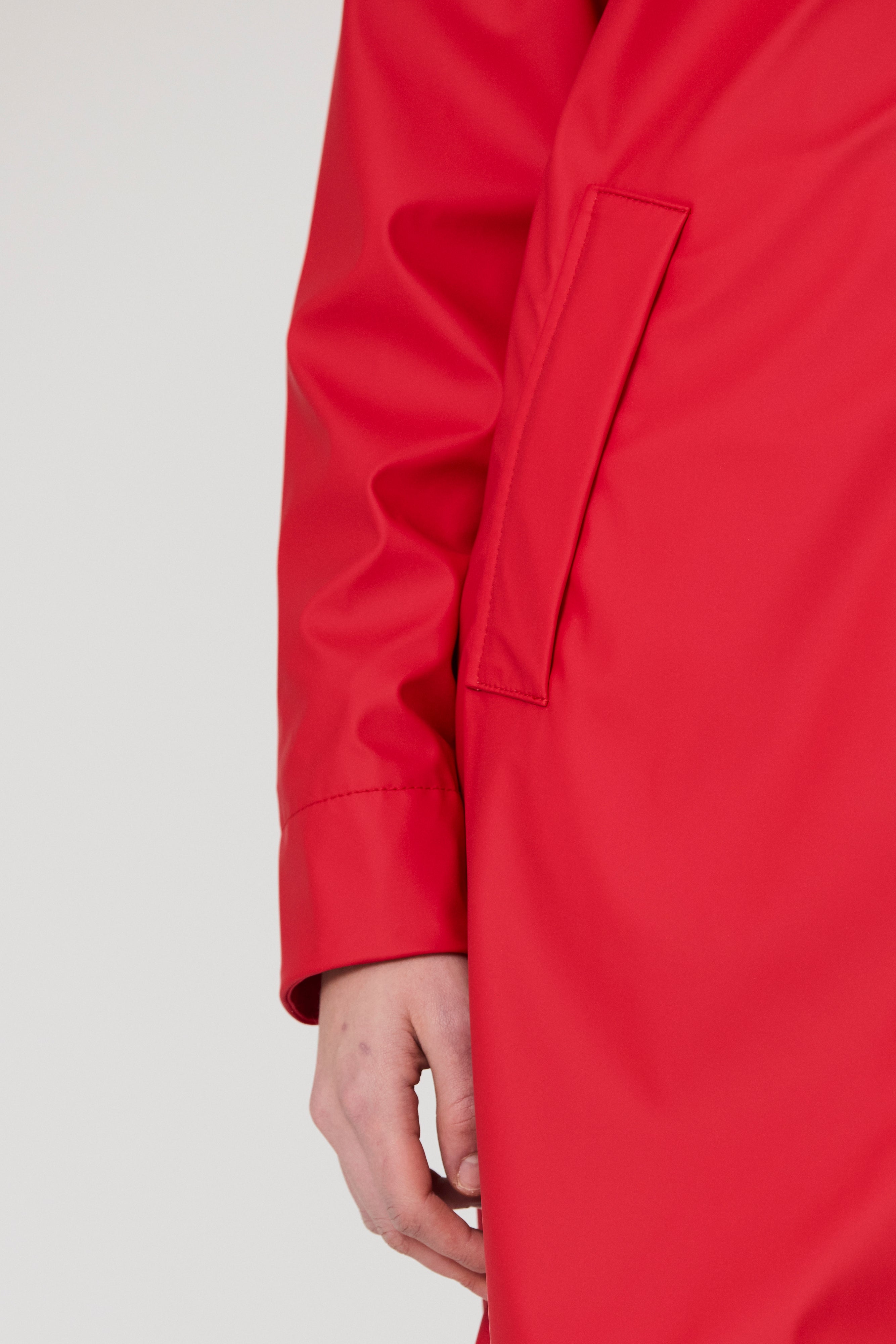 Close-up of a bright red coat or jacket sleeve and body with a side pocket, worn by a model against a light gray studio background.