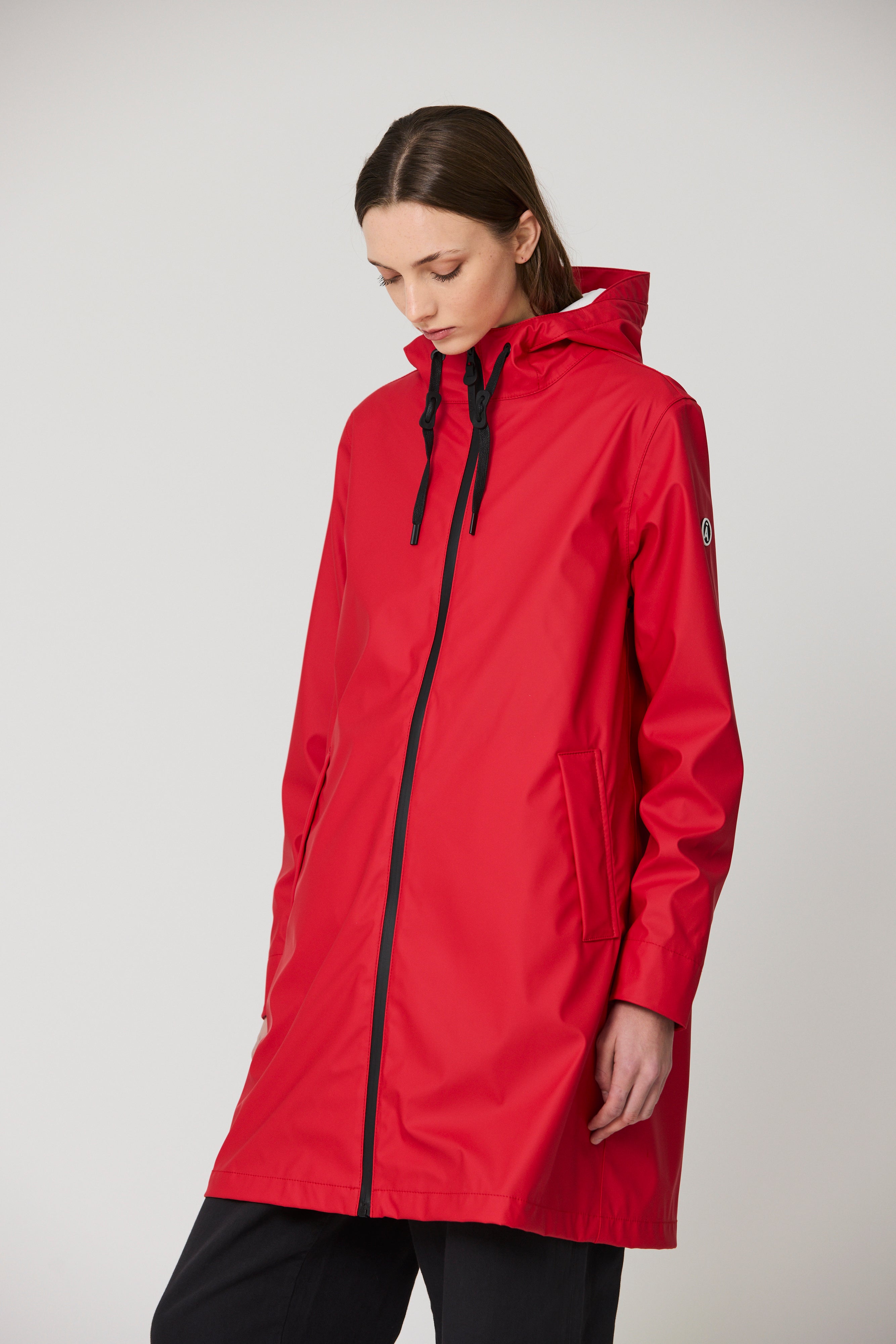 A woman wearing a bright red hooded raincoat with black drawstrings and a black front zipper, photographed against a plain light gray studio background.