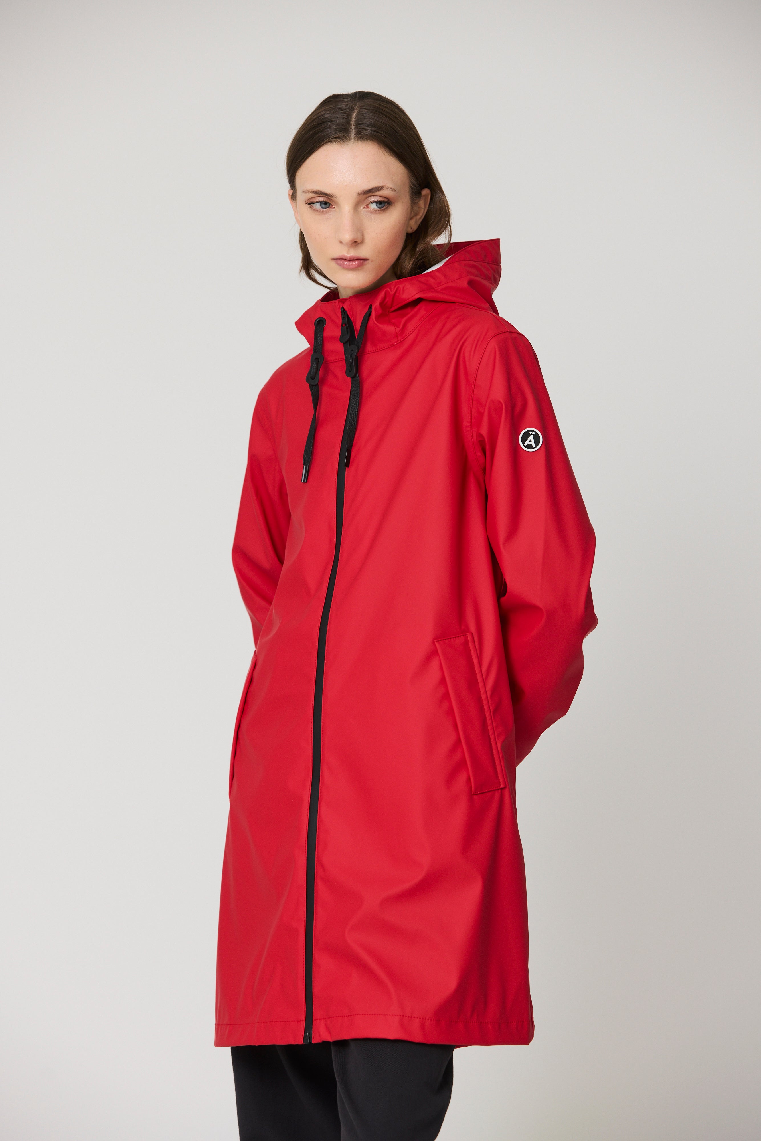 A woman modeling a bright red hooded raincoat with black zipper and drawstring details, shown against a plain light gray studio background.