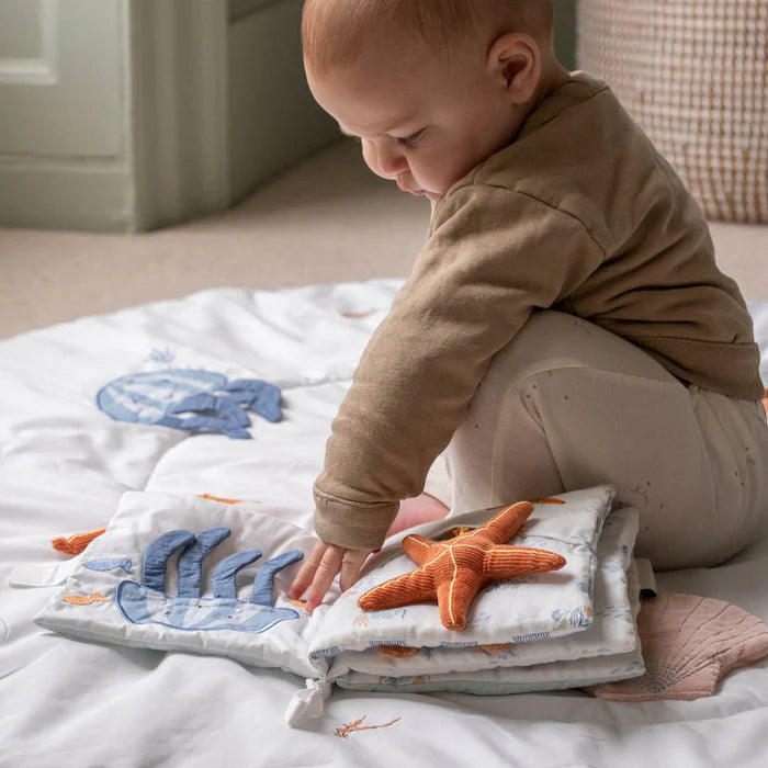 Baby playing with a soft book featuring marine animals on a bed.