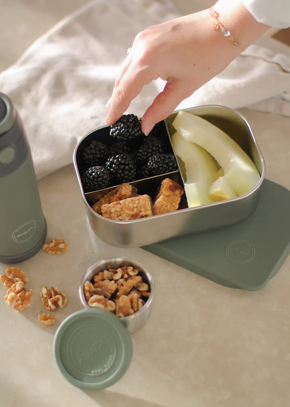 Hand reaching into a green bento box filled with snacks on a light surface.