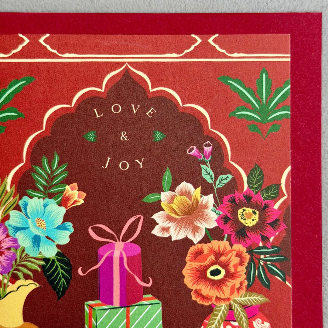 A close-up of a red decorative card or gift wrap featuring an ornate arch design, the words 'LOVE & JOY' in the center, and colorful illustrated flowers and gift boxes below.
