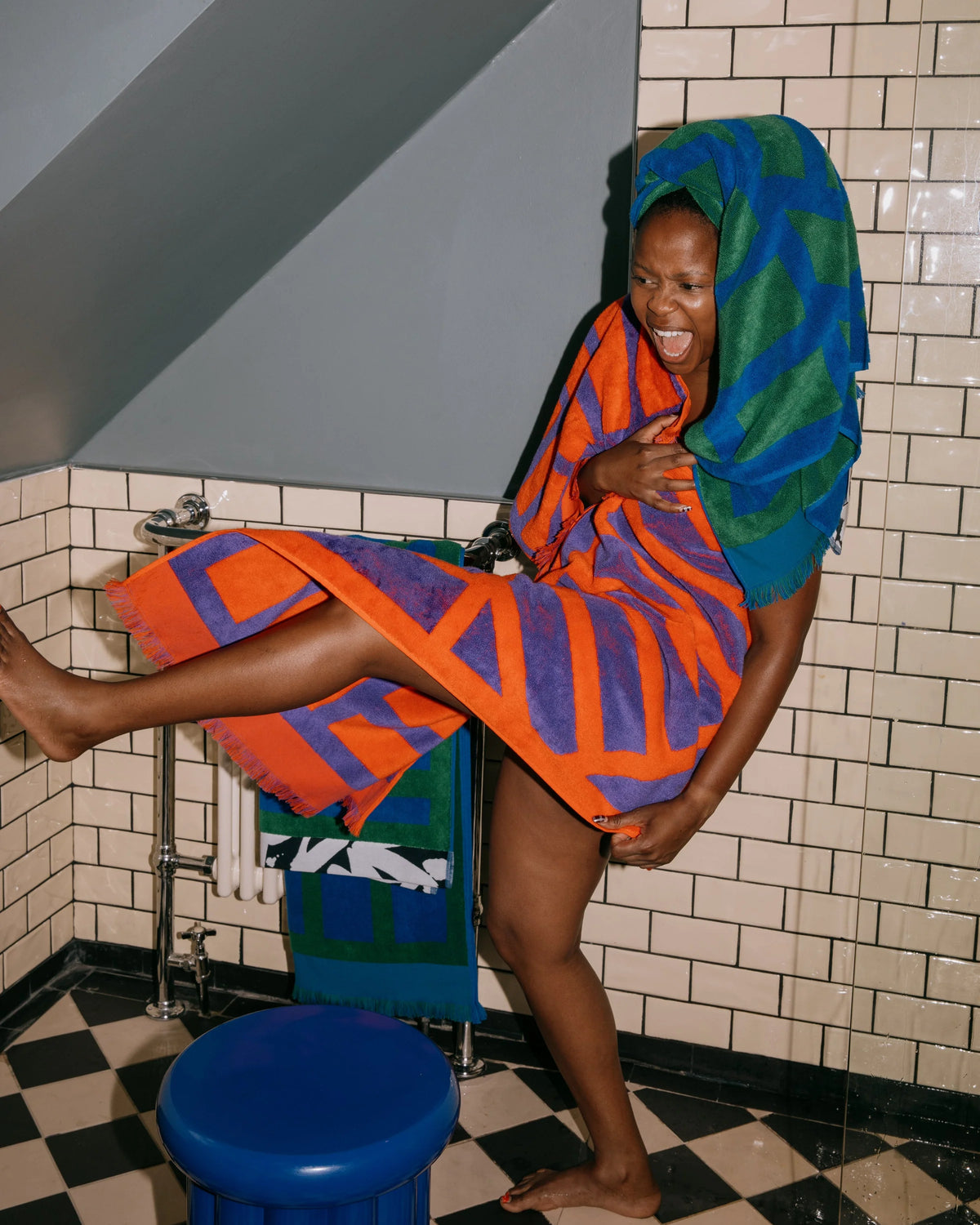 A woman wrapped in a bright orange and purple patterned towel sits on a blue stool in a tiled bathroom, with a matching towel draped over her head.