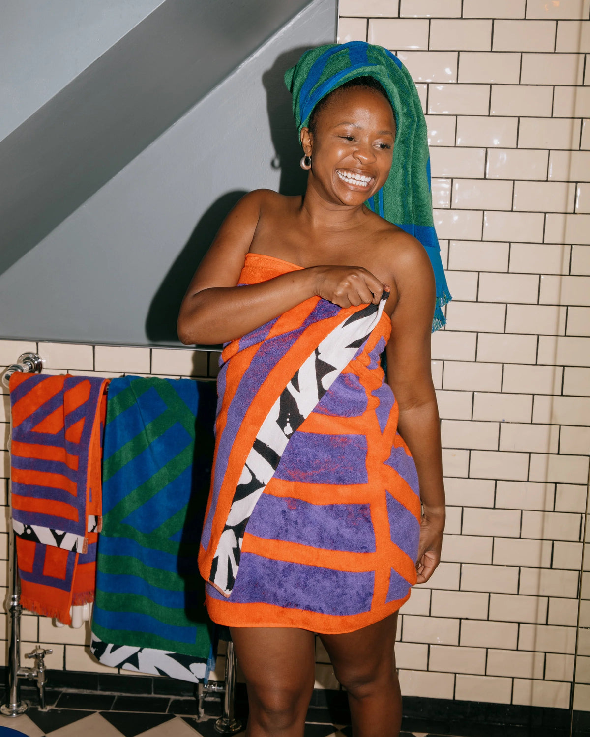 A smiling woman wrapped in a vibrant orange, purple, black, and white patterned towel, with a matching green and blue towel wrapped around her head, standing in a tiled bathroom next to a towel rack.