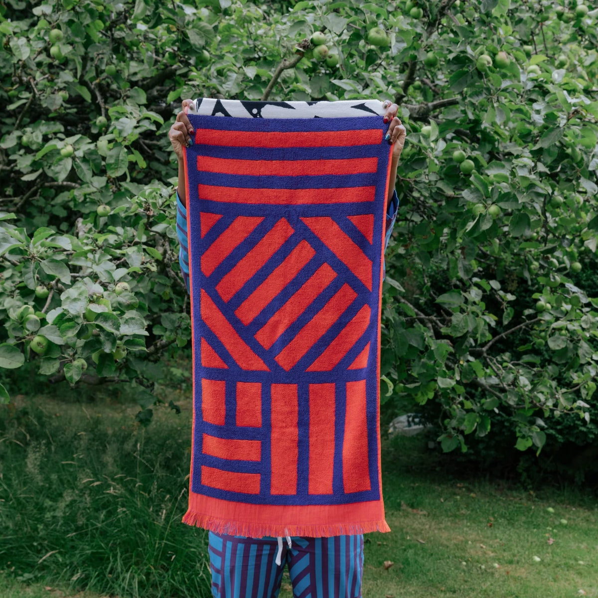 A person standing barefoot on grass in front of leafy green bushes, holding up a large woven textile with a bold geometric pattern in red and dark blue. The person is also wearing matching red and blue striped pants.