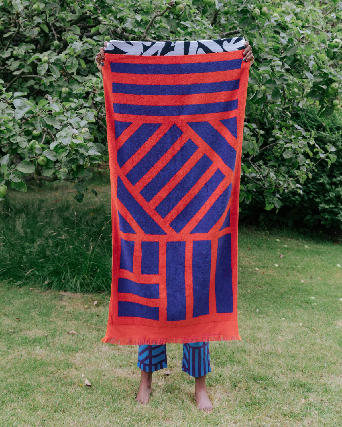 A person standing outdoors on grass in front of leafy bushes holds up a large rectangular textile with a bold geometric pattern in bright orange-red and deep blue. The person's legs and feet are visible below the cloth, wearing blue patterned pants.