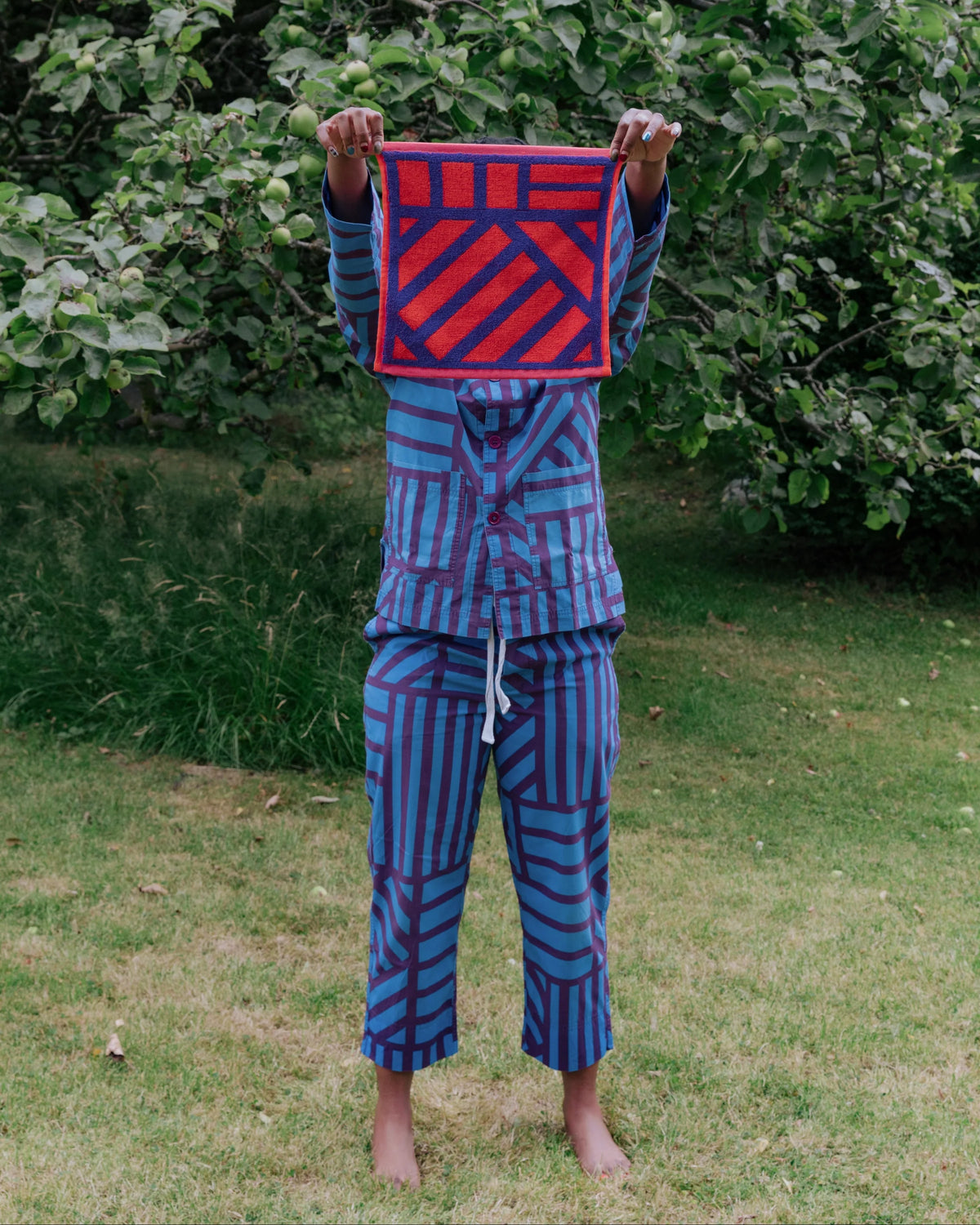 A person standing barefoot in a grassy outdoor area with leafy trees behind them, holding up a square red and blue patterned textile in front of their face. The person is also wearing matching blue and red patterned clothing.
