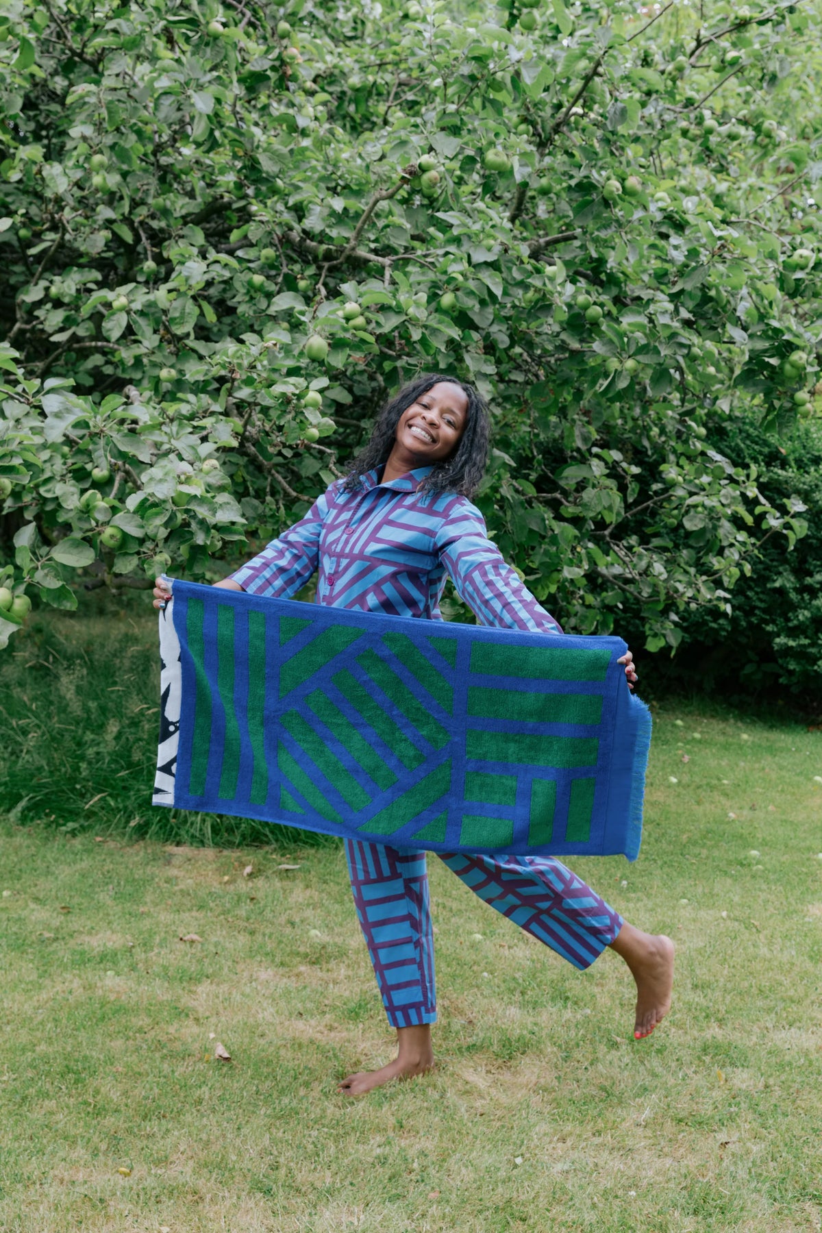 A woman stands barefoot on grass in front of leafy green bushes, holding up a large patterned textile. She is wearing matching blue and purple patterned pajamas or a top and pants set.