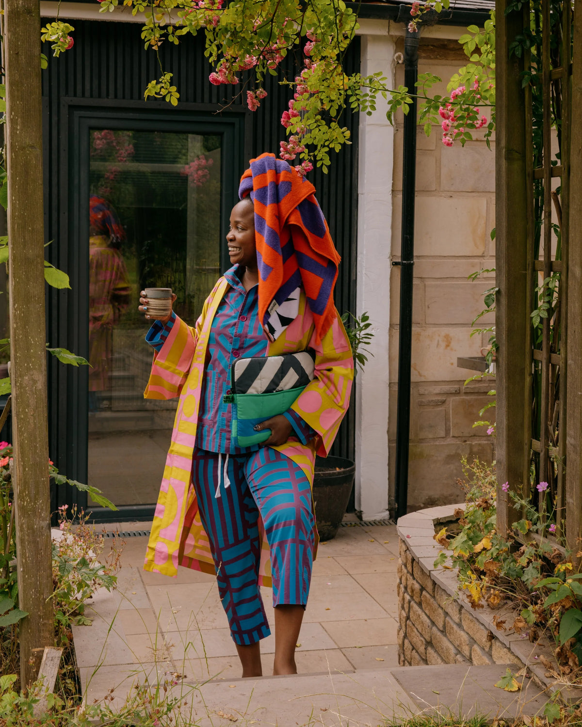 A woman stands in a garden courtyard under a wooden arbor, wearing a vibrant multicolor patterned outfit with a matching head wrap. She holds a glass and a green bag, with a stone building and climbing plants in the background.