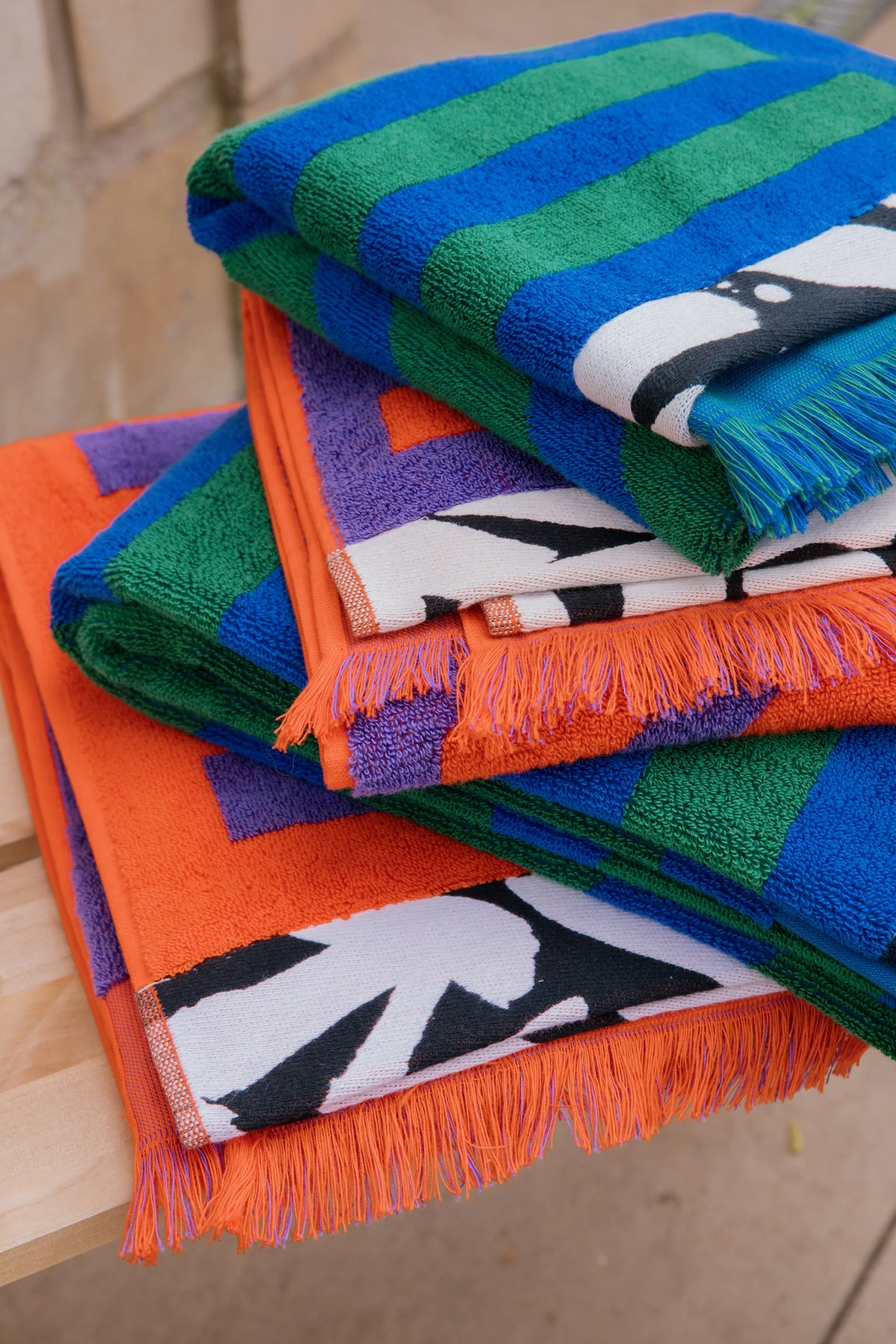 A stack of colorful striped and patterned towels with fringed edges, including blue, green, orange, purple, and white designs, displayed on a wooden surface.