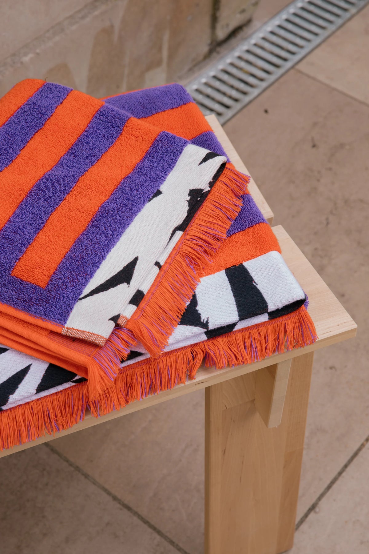 A folded multicolor striped towel with orange, purple, white, and black patterns and fringed edges, displayed on a light wood table.