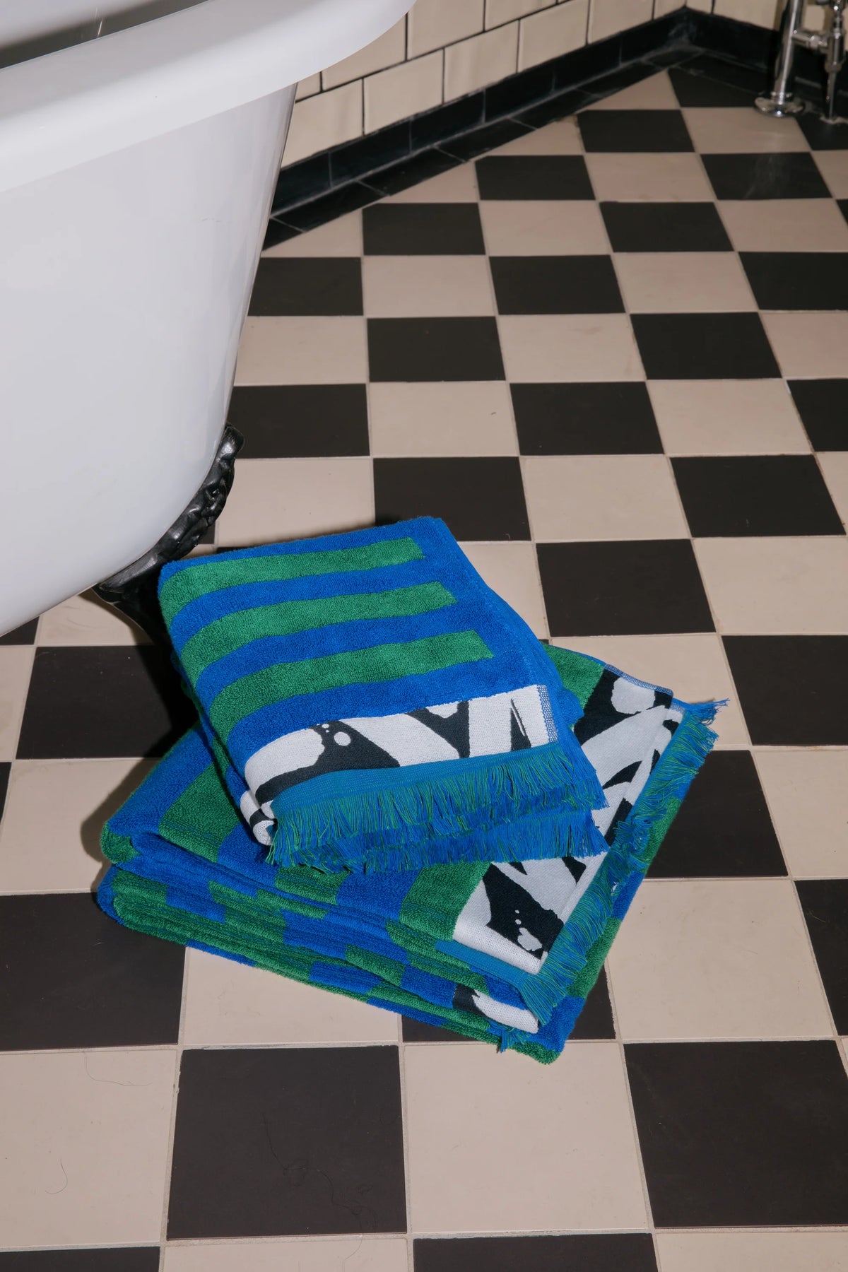 A stack of folded bath towels in a bathroom setting on a black and white checkered floor, next to a white bathtub. The towels feature blue and green stripes, and one towel displays a black and white penguin print.