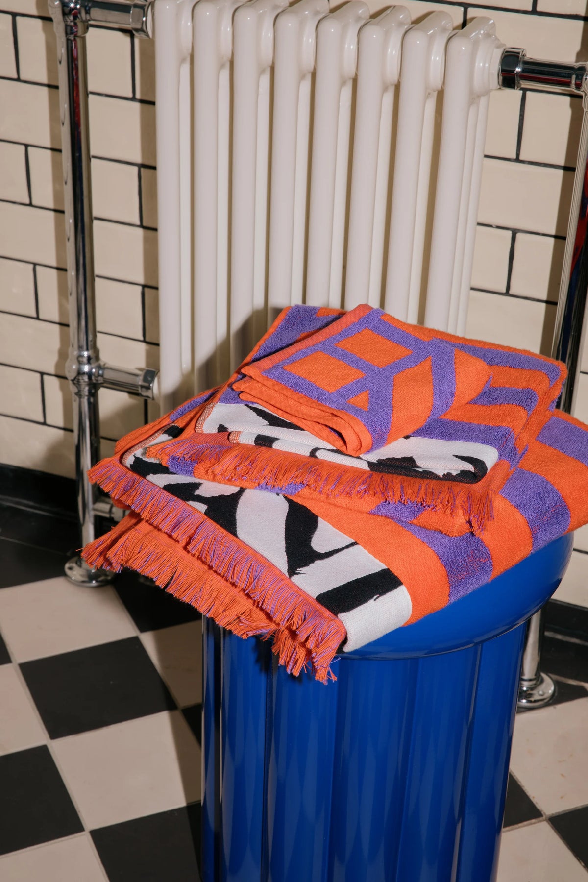 A folded orange, purple, black, and white patterned fringed textile, likely a throw blanket or scarf, displayed on top of a blue cylindrical container in front of a white radiator and tiled wall.